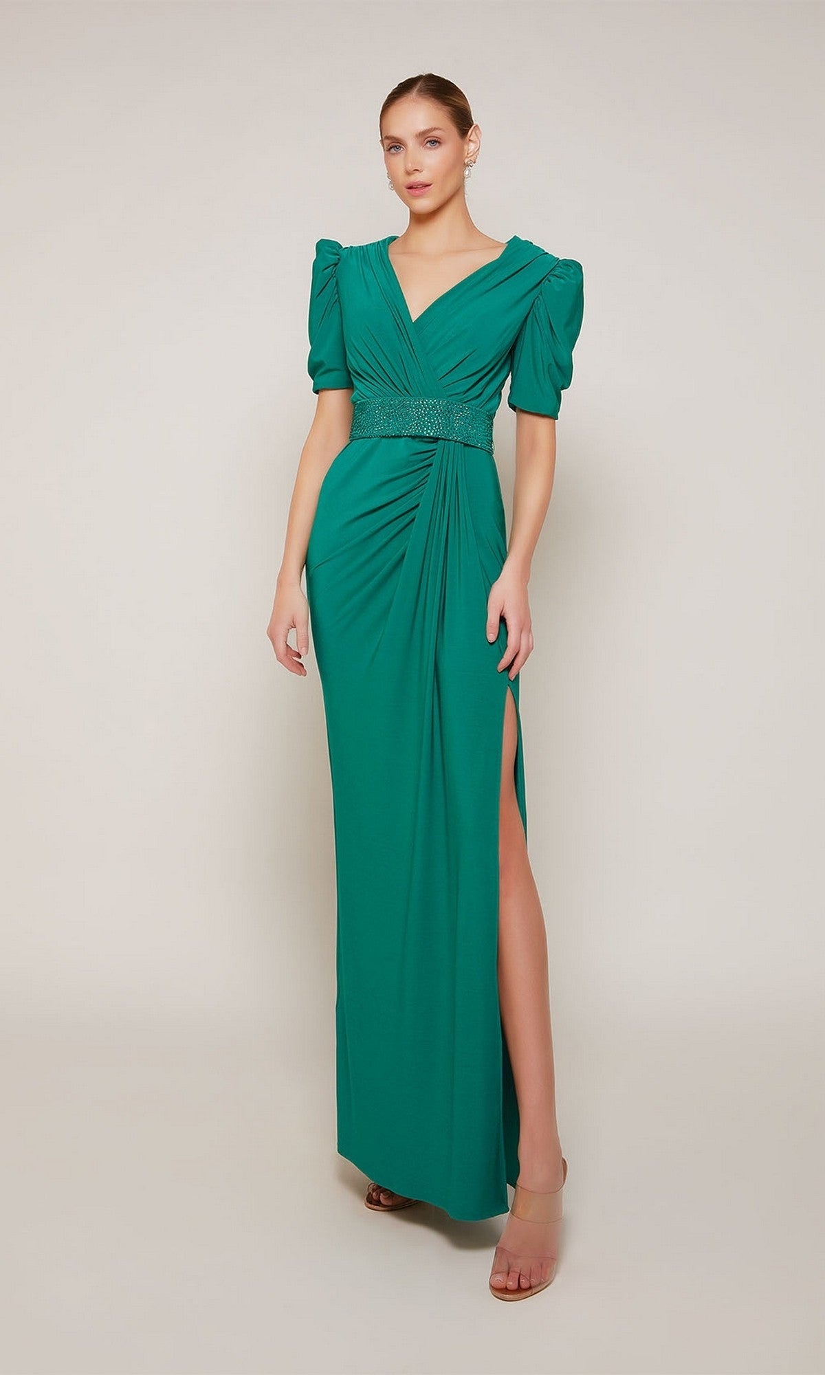 A woman wears the Alyce 27690 long formal dress in emerald, featuring a v-neckline, short puffed sleeves, gathered waist, and thigh-high slit, paired with nude heels against a plain background.
