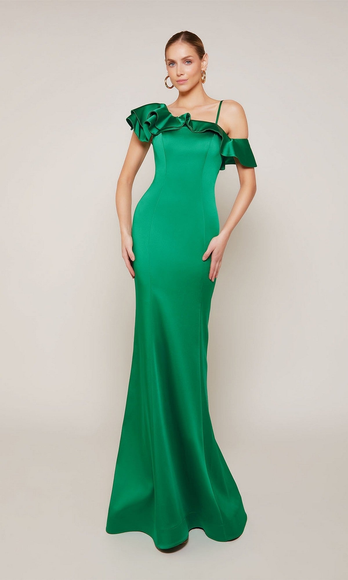 A woman stands against a plain light background wearing the Alyce 27691 long formal dress in emerald satin, featuring a fitted silhouette, one-shoulder ruffle detailing, and gold hoop earrings.