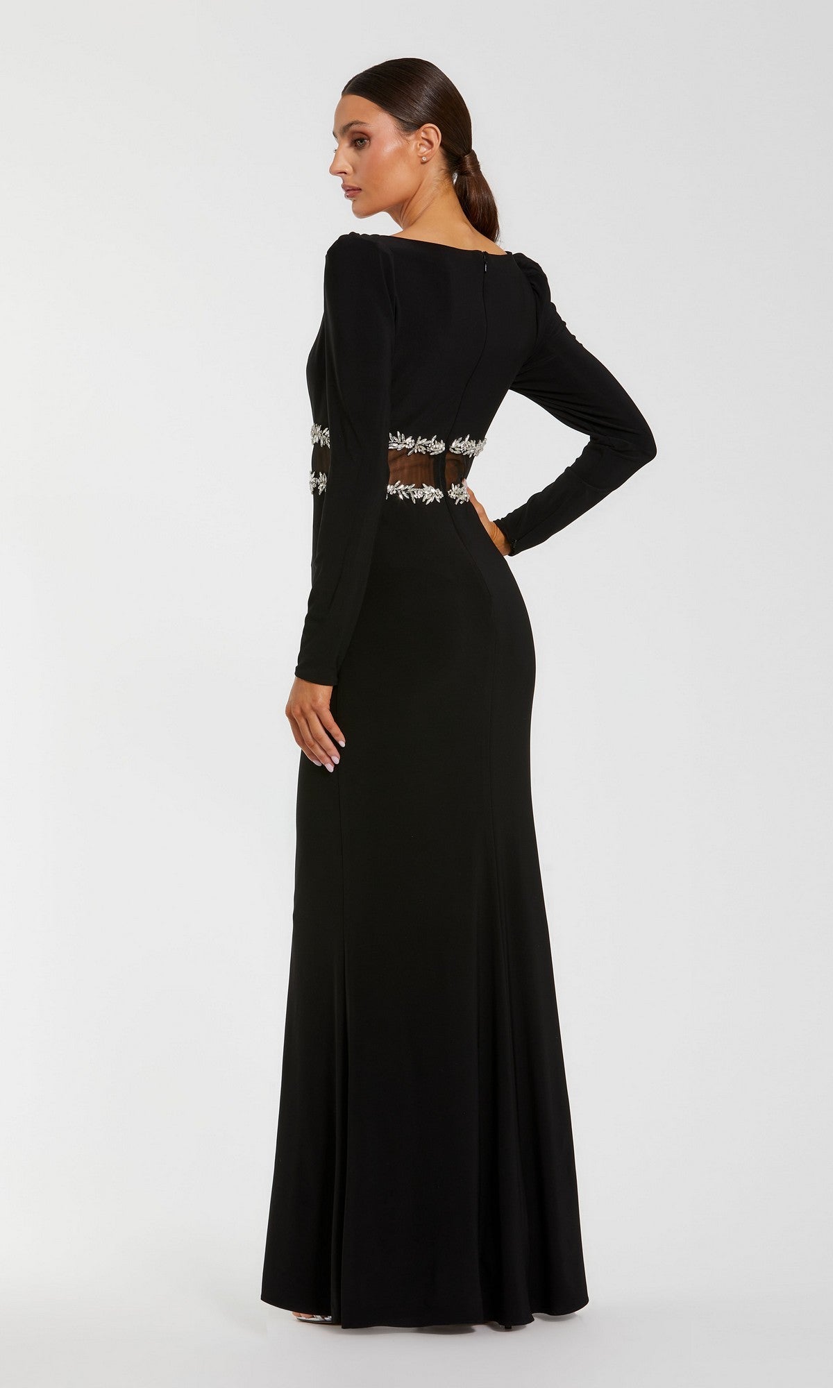 A woman is wearing the Mac Duggal 27695, a long-sleeved, floor-length black formal evening gown featuring puff shoulders and a sheer embroidered waist detail, crafted from stretch jersey fabric, while standing against a plain white background.