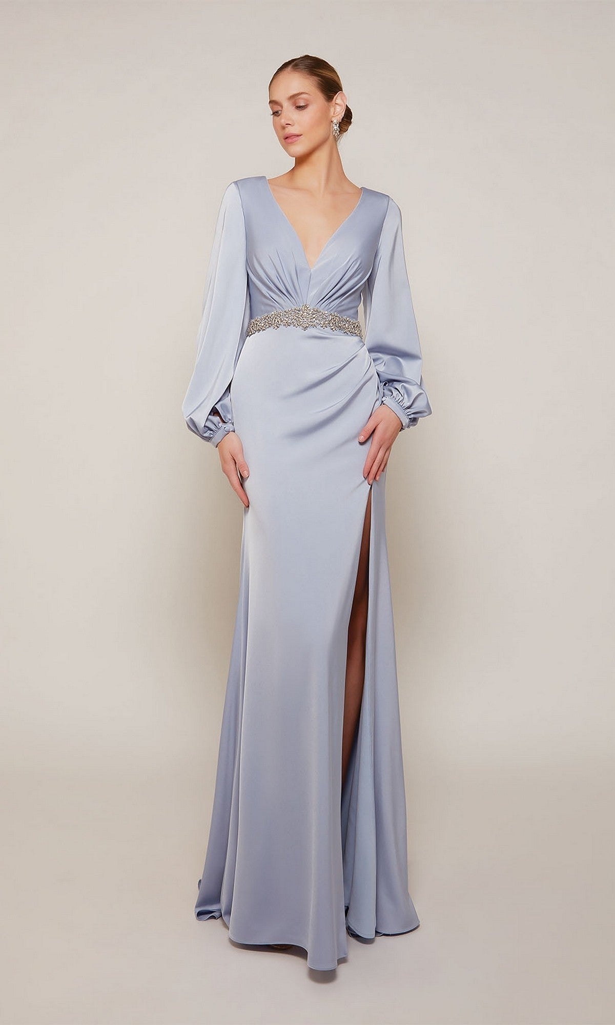A woman wears the Alyce 27696 long formal dress in French Blue, featuring long sleeves, a deep V-neckline, jeweled waistband, and high slit, standing against a plain background.