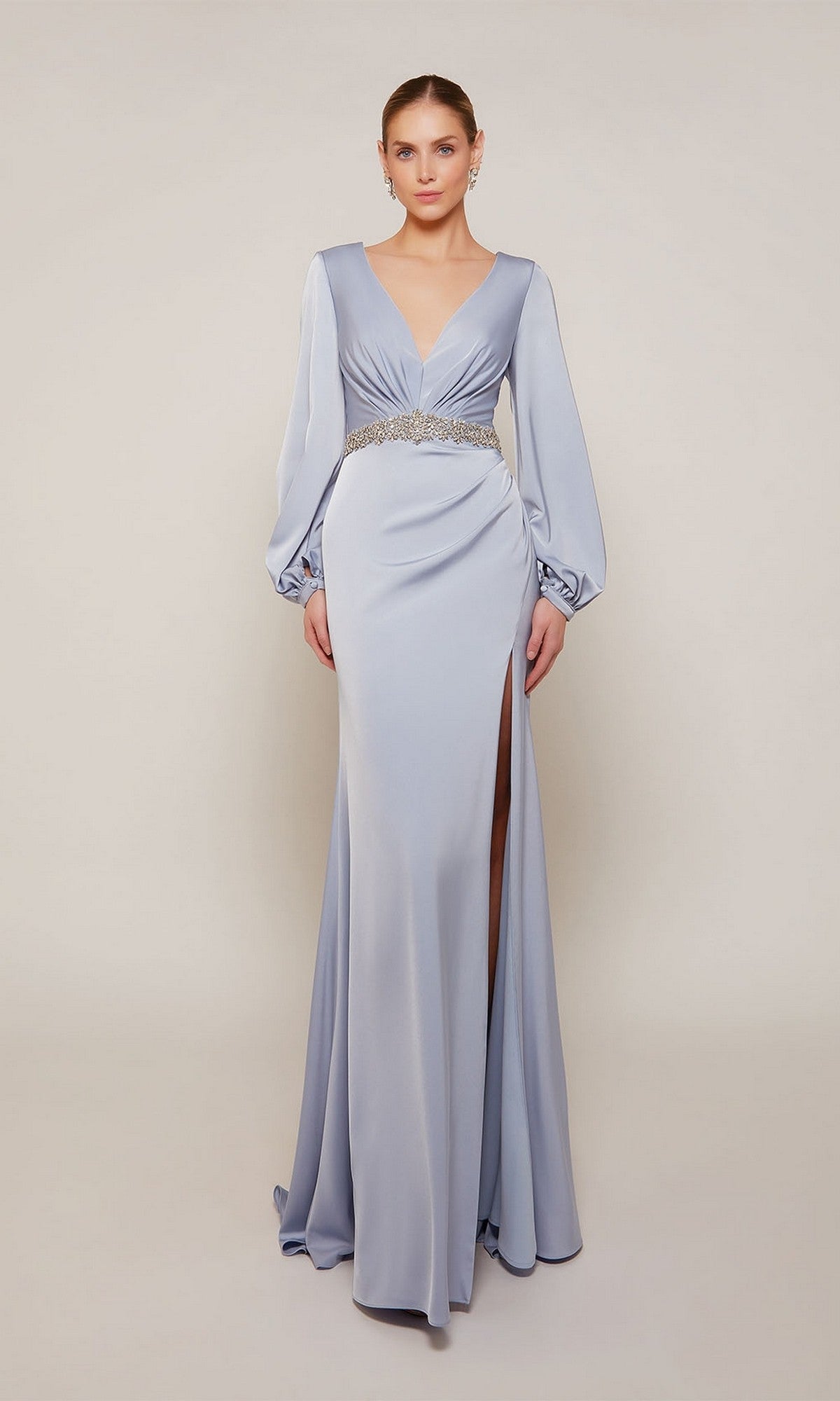 A woman models the Alyce 27696, a long, light blue satin formal dress featuring long sleeves, a deep V-neckline, jeweled waist detail, and a high slit, standing against a plain backdrop.