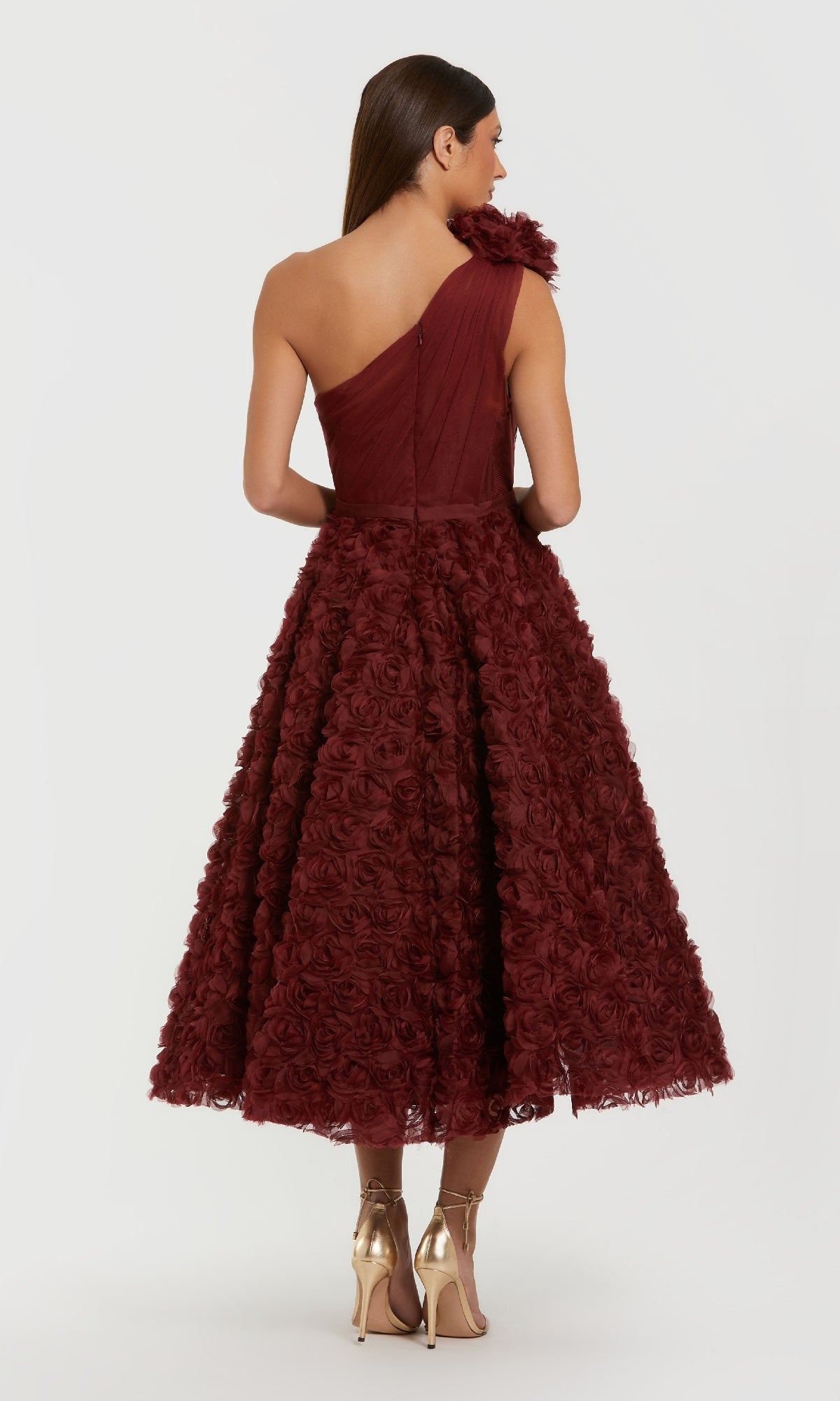 A woman faces away, wearing the Mac Duggal 27698 Midi Wine Wedding Guest Dress with a textured floral skirt and gold heels, set against a plain white background.