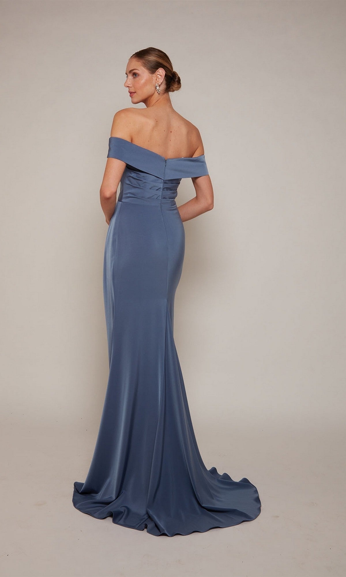 A woman in the Alyce 27698 long formal dress—a fitted, off-the-shoulder blue gown—stands facing away with her hands behind her back against a plain background.