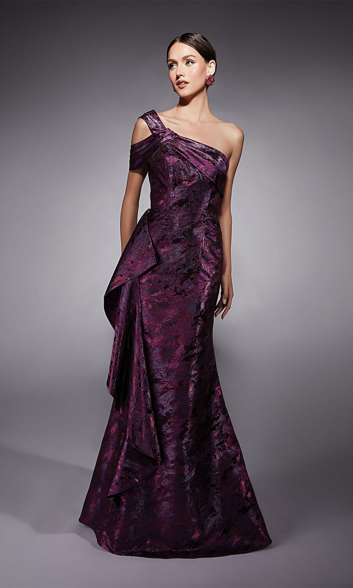 Woman wearing an Alyce 27701 long formal dress in fuchsia, featuring a floor-length, one-shoulder jacquard design with a textured metallic finish, standing against a plain gray background.