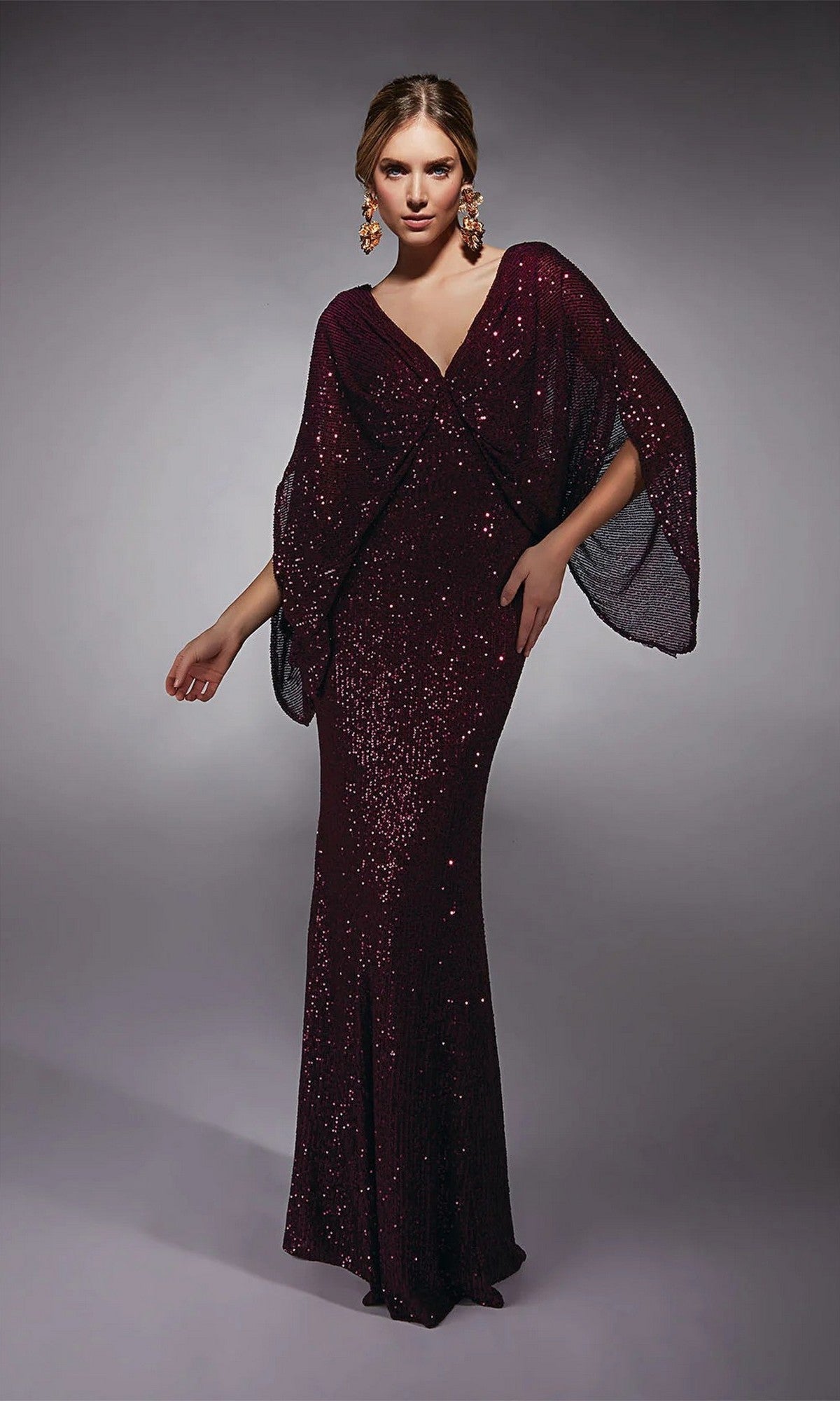 A woman models the Alyce 27703 Long Formal Dress in aubergine, featuring floor-length sequins and wide draped sleeves. Paired with statement earrings, it's ideal as a mother-of-the-bride or formal evening gown against a gray backdrop.