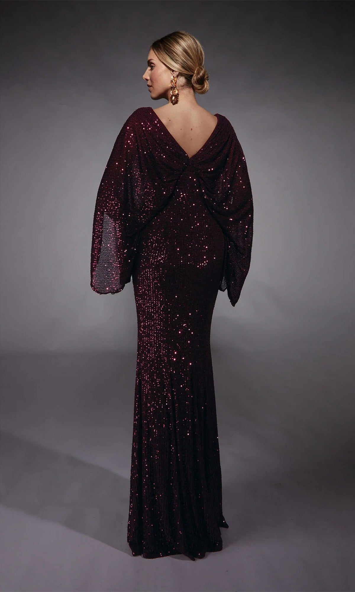A woman stands with her back to the camera, showcasing the Alyce 27703— a floor-length, dark burgundy sequin gown with draped sleeves and a low back, perfect for mothers of the bride or any formal evening event.