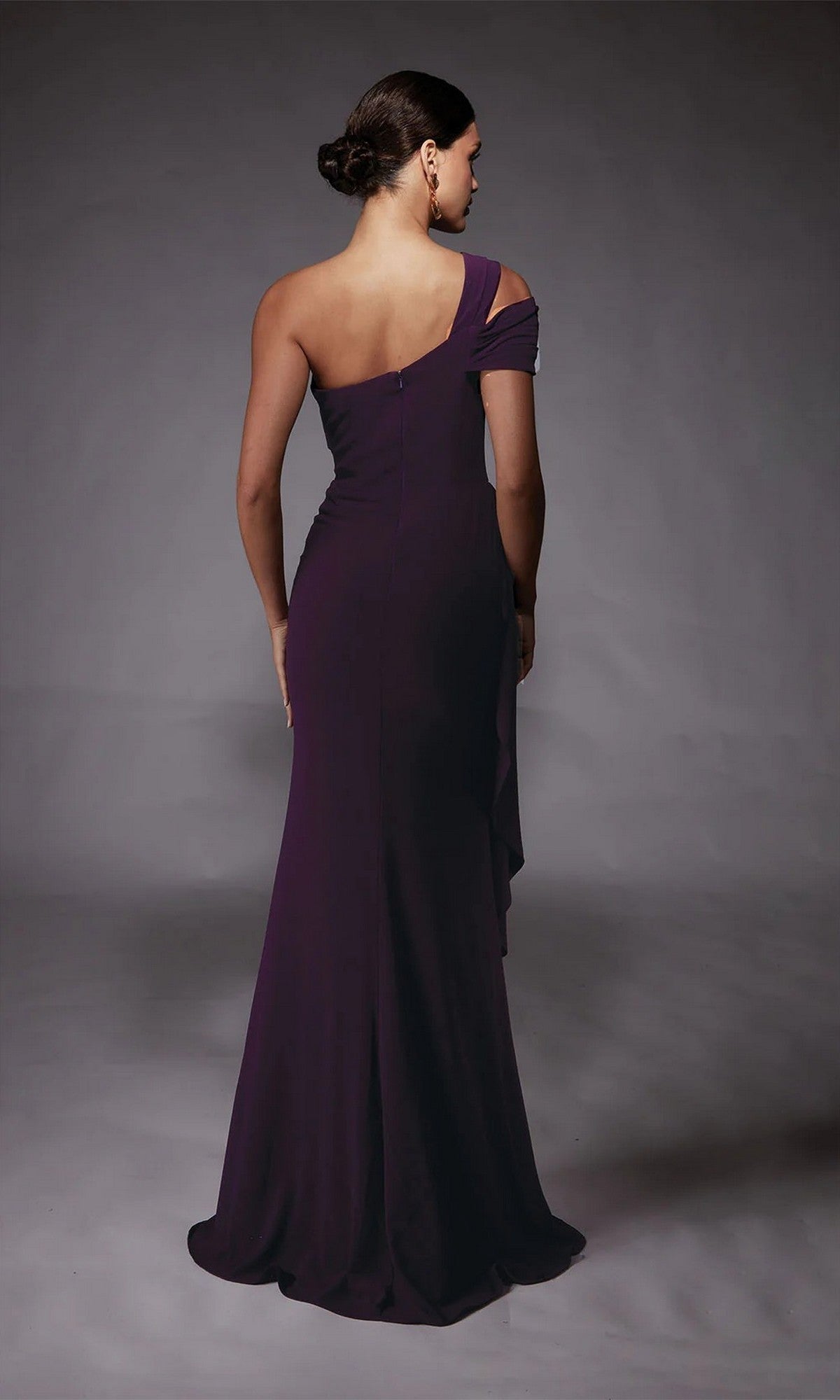 A woman with a low bun faces away, modeling the Alyce 27705 long formal dress—floor-length, one-shoulder, and dark purple—set against a plain gray background.