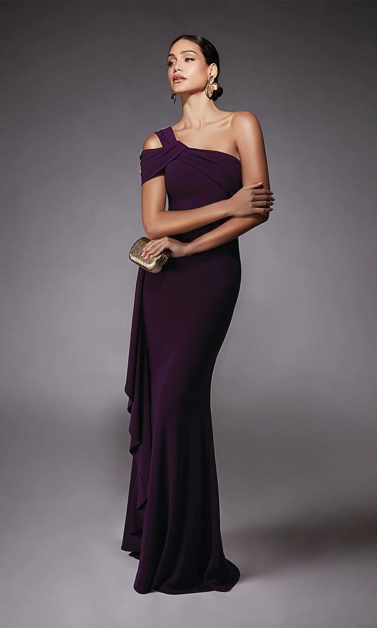 A woman wears the Alyce 27705 long formal dress with cold shoulder sleeves, holding a small gold clutch, standing against a plain gray background.