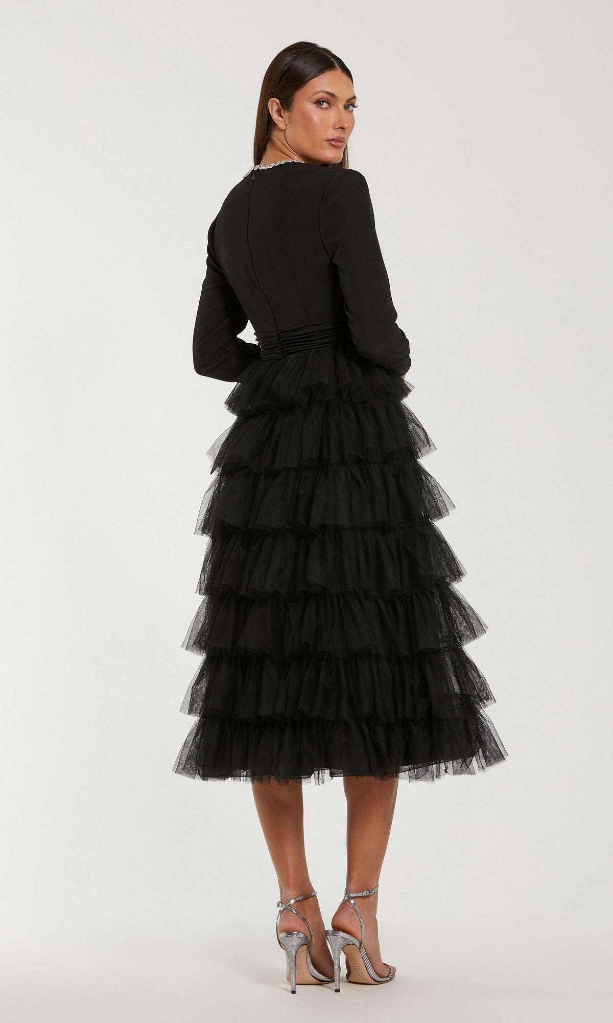 A woman stands facing away from the camera, wearing the Midi Wedding Guest Dress: Mac Duggal 27707—a black midi dress with a tiered tulle skirt—paired with silver high heels on a plain white background, making it an elegant and modest choice for a wedding guest outfit.