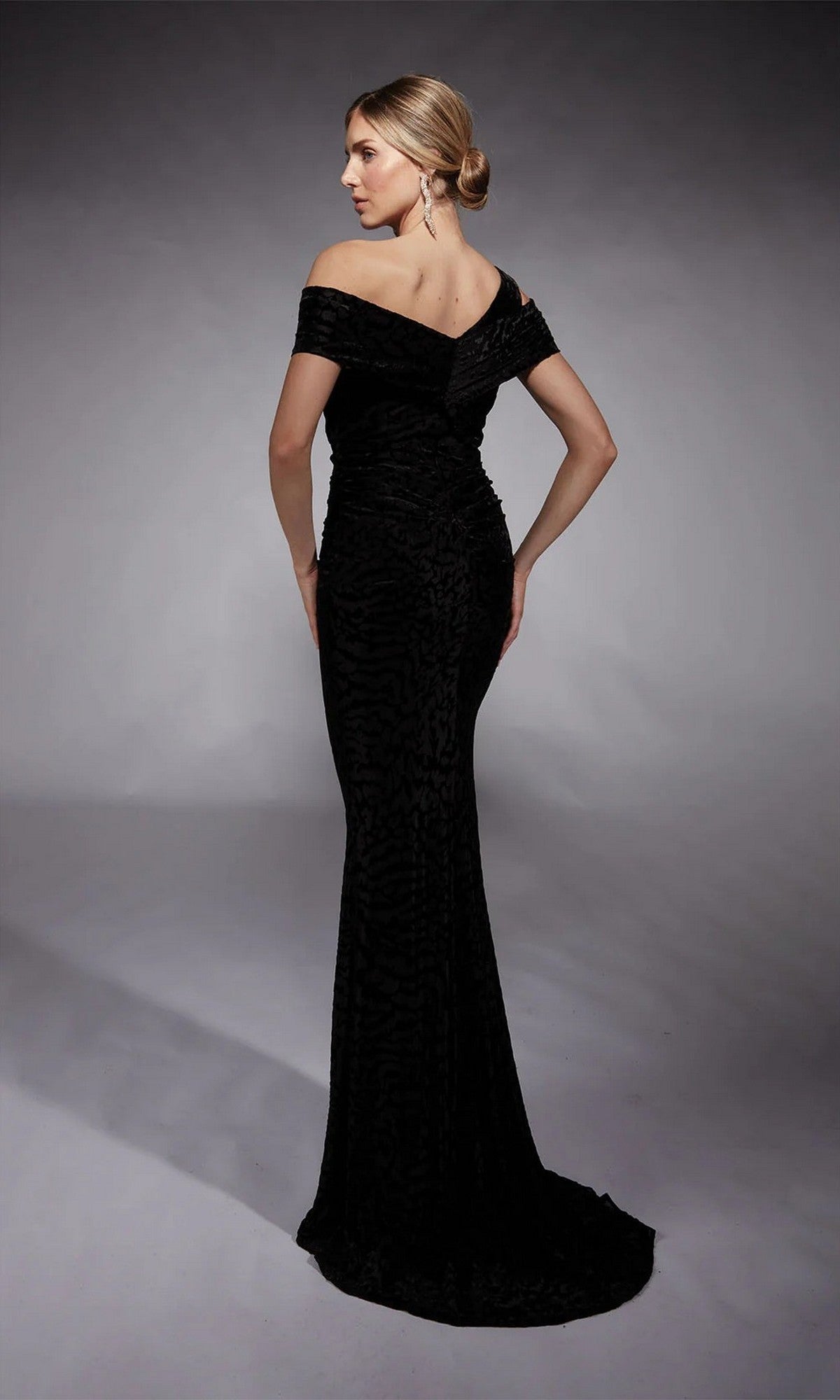 In a studio setting, a person faces away wearing the Long Formal Dress: Alyce 27709, which features an off-the-shoulder asymmetric neckline, fitted silhouette, and textured pattern.