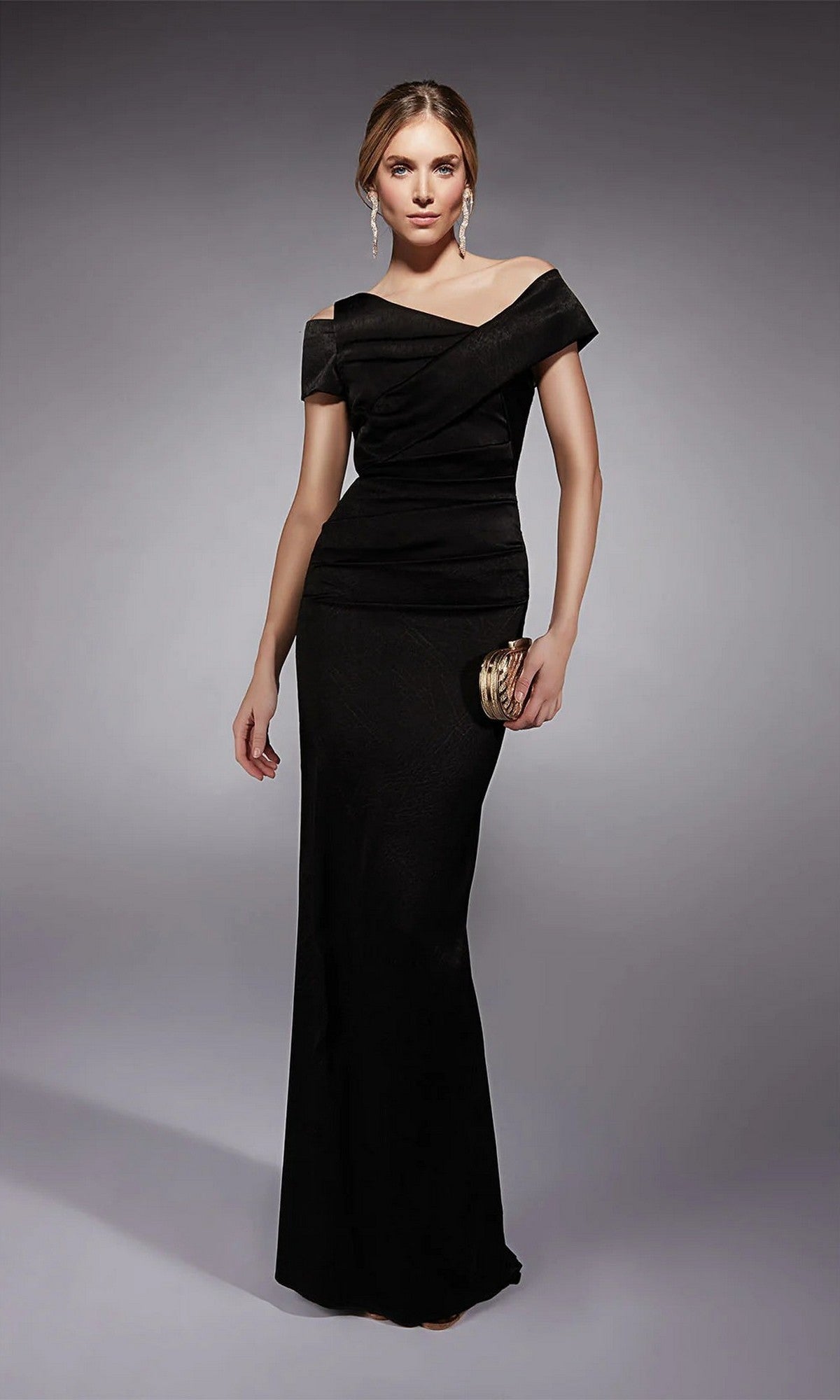 A woman stands in a studio wearing the black Alyce 27710 long formal dress, holding a small gold clutch, with her hair styled back and minimal accessories.