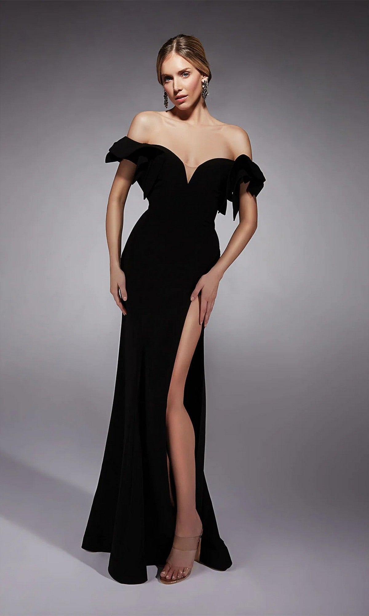 A woman stands against a plain, light-gray background wearing the Alyce 27713 long formal dress in black, featuring an off-shoulder design with a high slit.