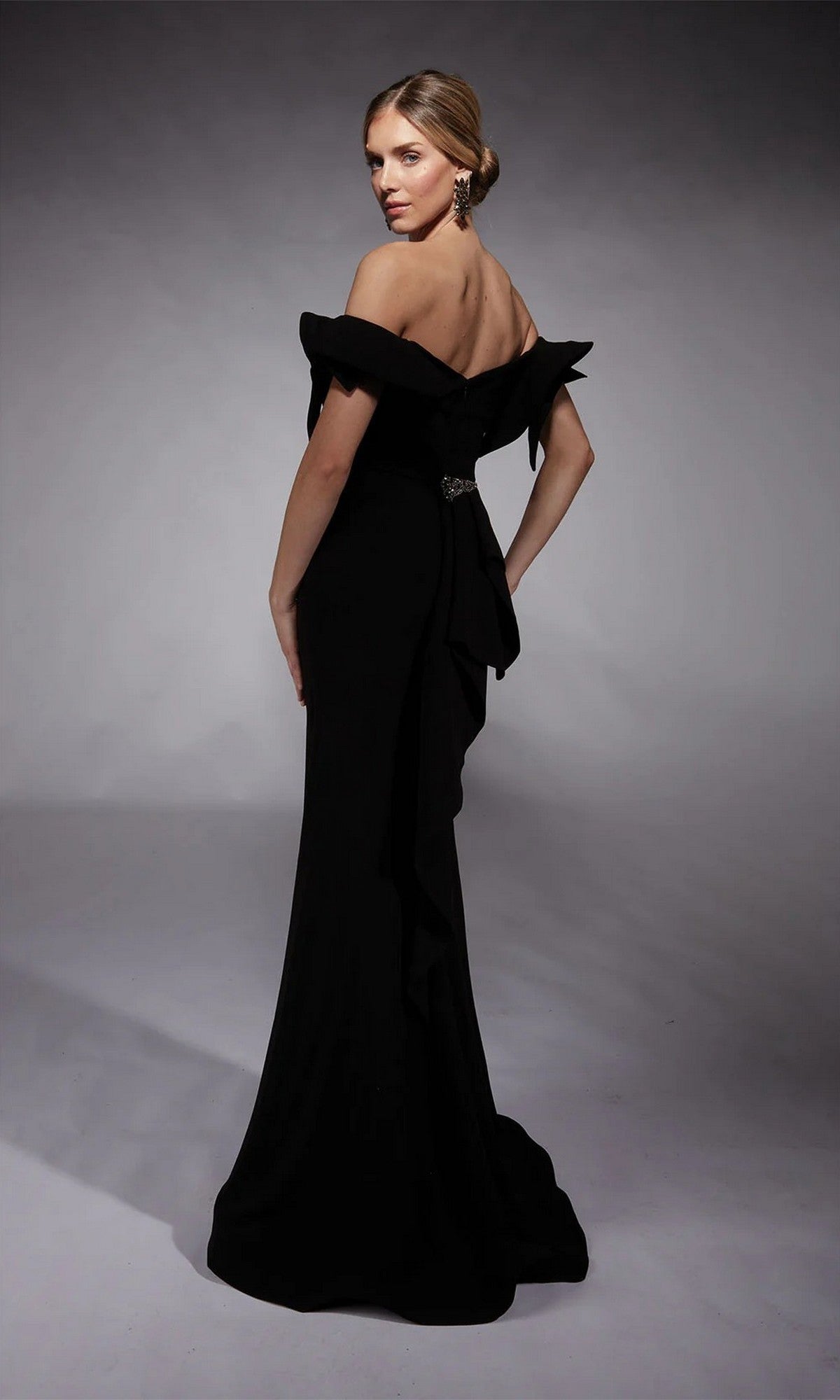 A woman models the Alyce 27713 long formal dress—an off-shoulder, floor-length black gown with ruffle sleeves—standing on a gray studio backdrop and looking over her shoulder with her hair in a low bun.