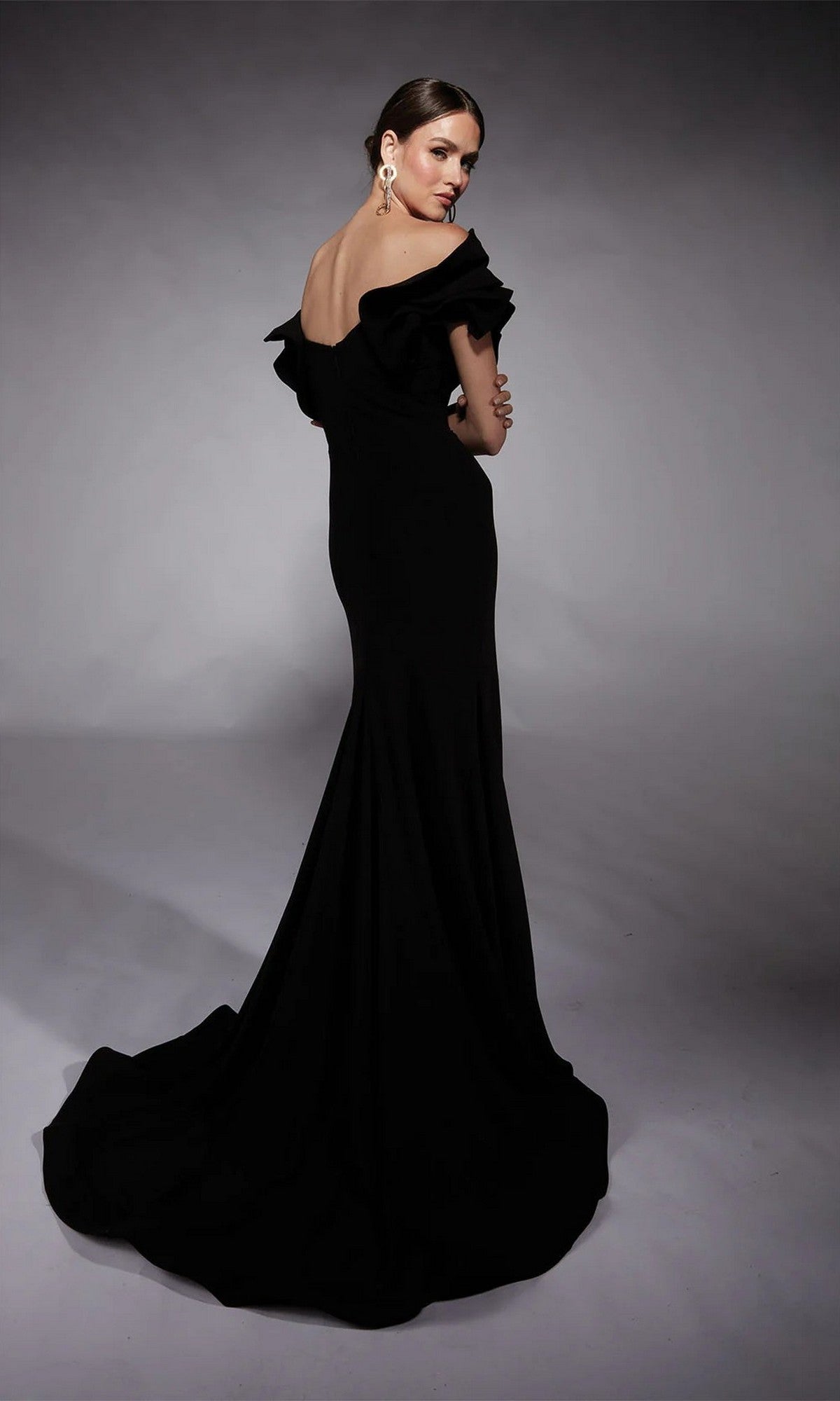 A woman stands against a plain gray backdrop, looking over her shoulder in the Alyce 27714 long formal dress—an elegant off-shoulder black gown with a sheath silhouette and sweeping train.