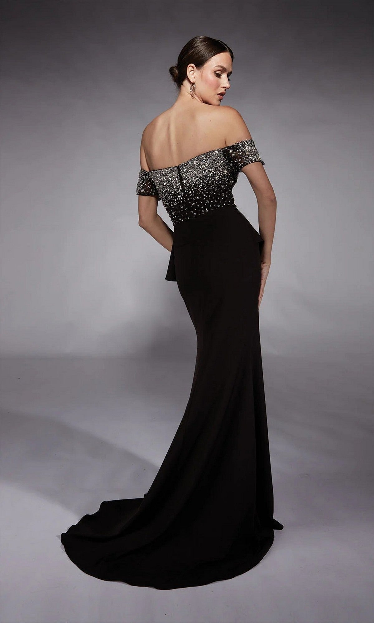 A woman models the Alyce 27715 long formal dress—an elegant black off-the-shoulder gown with a fitted beaded peplum bodice—standing with her back turned and head slightly tilted.