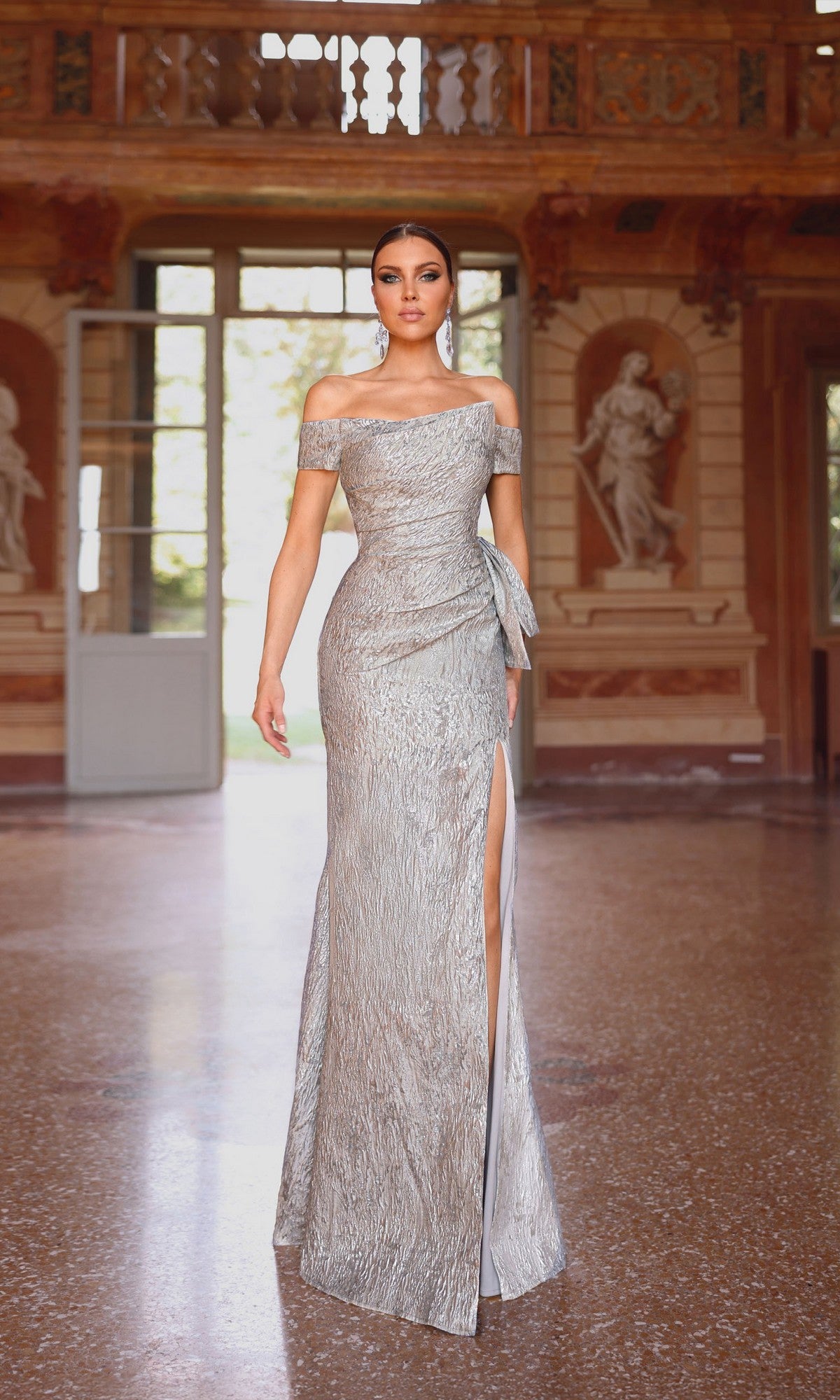 A woman wears the Alyce 27717 long formal dress, featuring textured jacquard fabric, an asymmetrical neckline, and a thigh-high slit, standing in an ornate room with classical statues and open doors in the background.