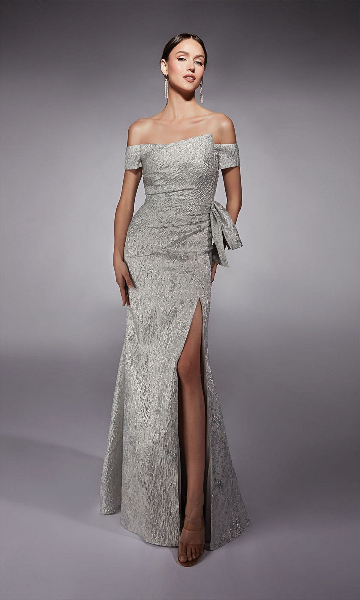 A woman models the silver Long Formal Dress: Alyce 27717, featuring textured jacquard fabric, an asymmetrical neckline, a thigh-high side slit, and a side bow detail, set against a plain gray background.