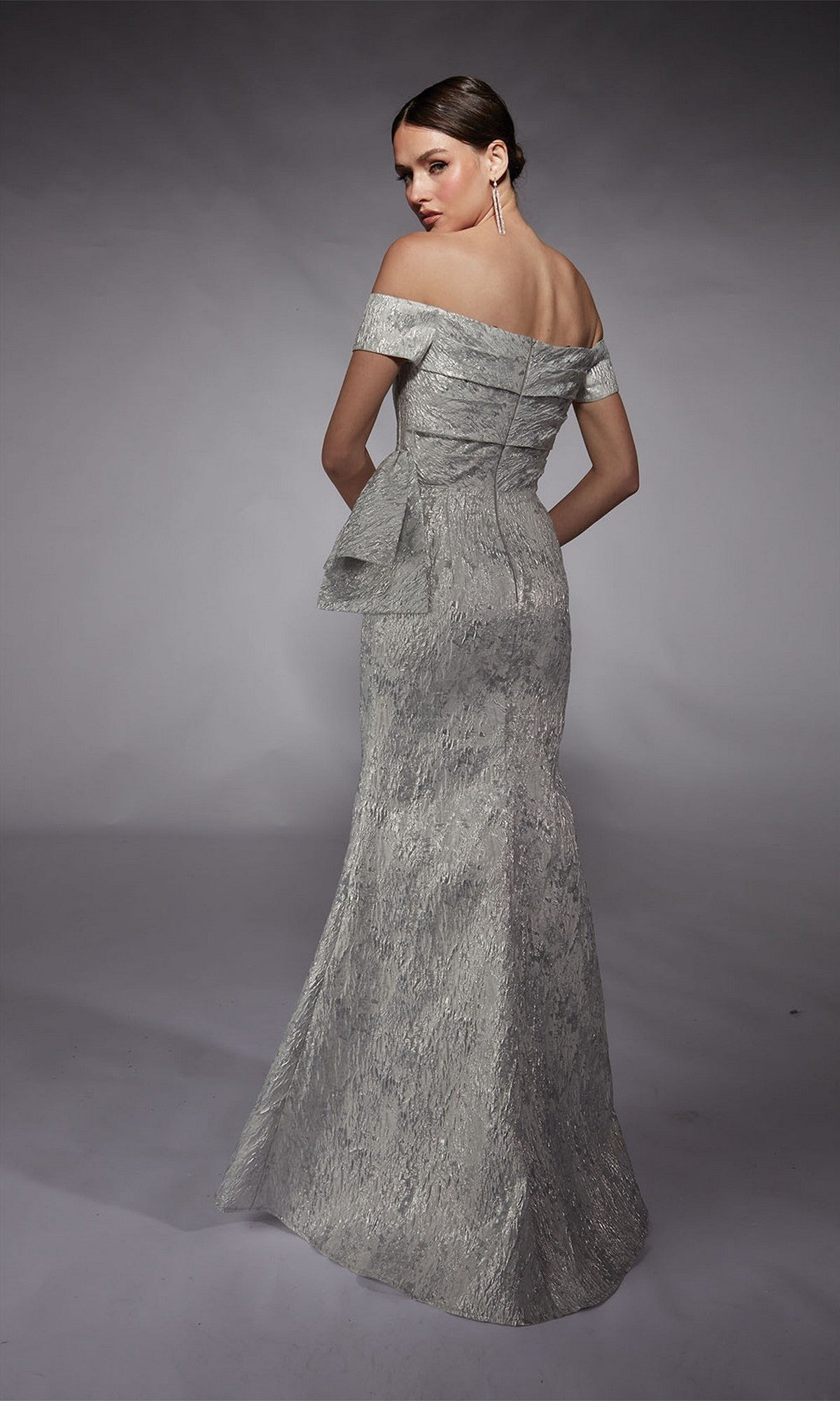A woman models the Alyce 27717, an off-the-shoulder, floor-length jacquard formal dress with a side bow detail, seen from behind against a plain studio backdrop.