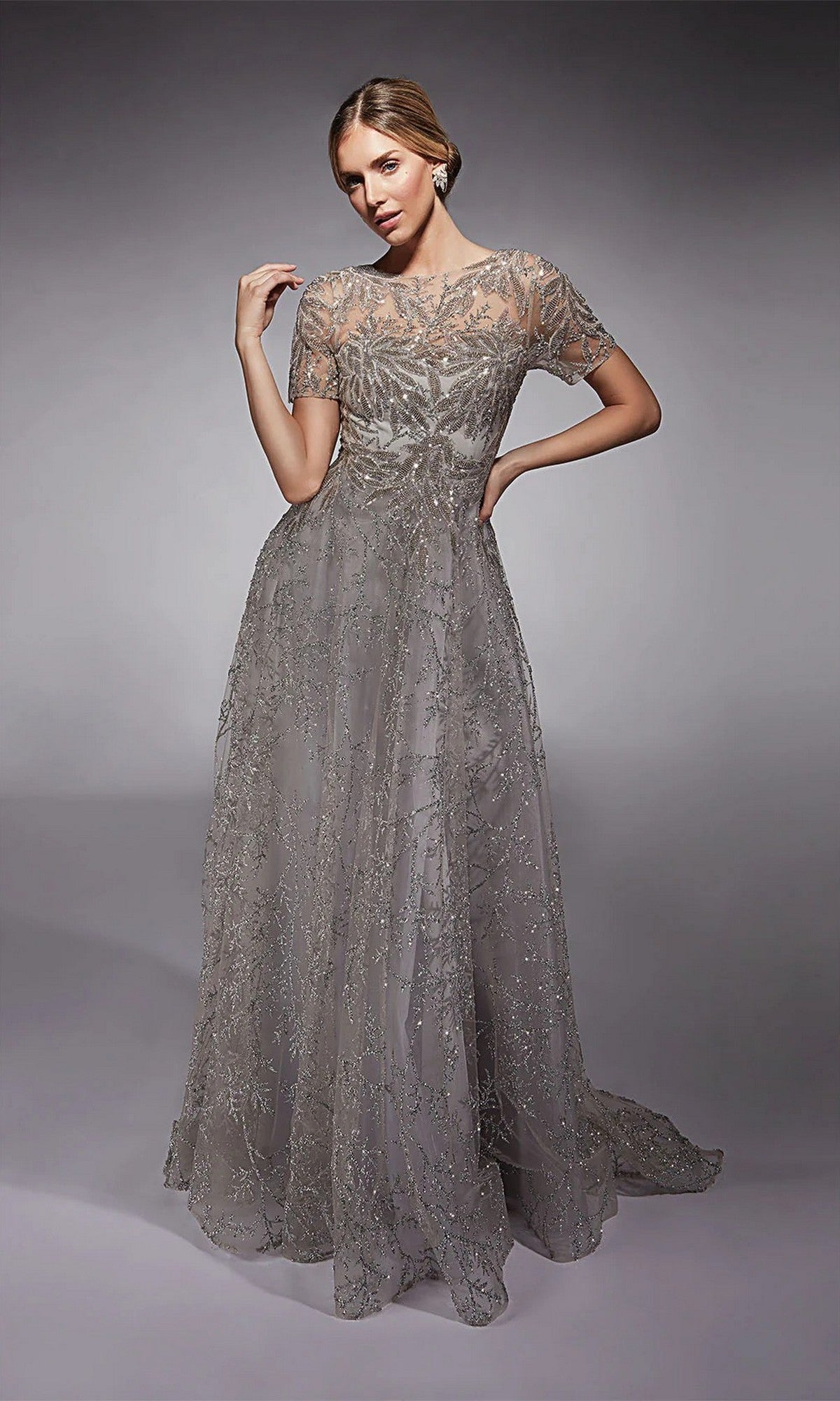 A woman poses against a plain gray background wearing the Alyce 27726 long formal dress—floor-length with short sleeves, intricate beaded embellishments, and a sheer glitter tulle overlay.