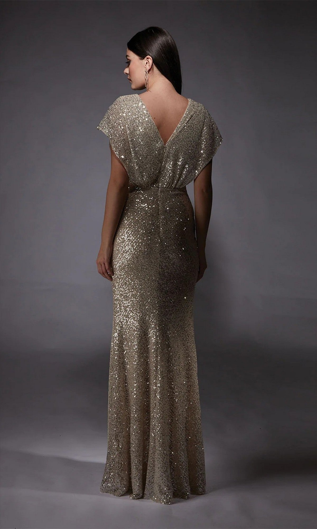 A woman faces away in the Alyce 27729 long formal dress, featuring floor-length gold sequins, short sleeves, and a deep V-back against a plain dark background.