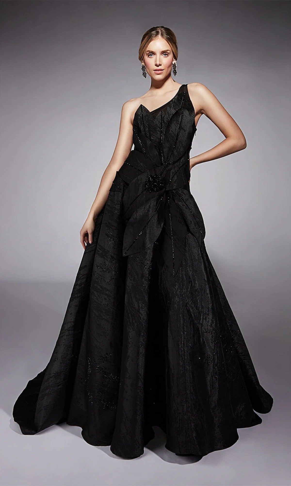 A woman in a black, sleeveless, floor-length Alyce 27730 jacquard formal dress with a textured fabric and asymmetric neckline poses with one hand on her hip against a plain gray backdrop—perfect for mother-of-the-bride events.
