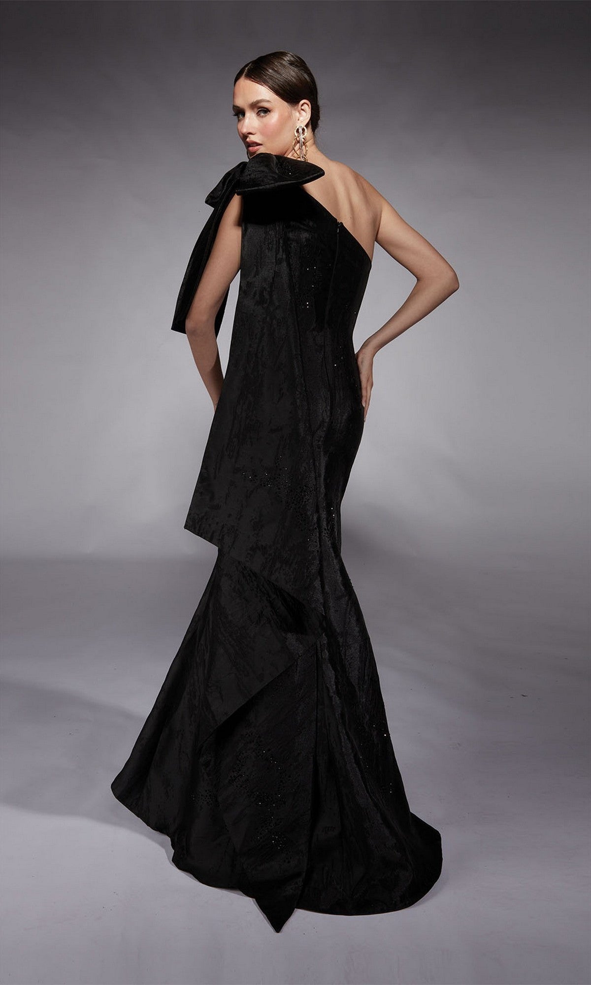 A woman models the Alyce 27731 long formal dress, featuring a one-shoulder design, dramatic bow detail, and flared skirt, as she poses against a plain gray background.