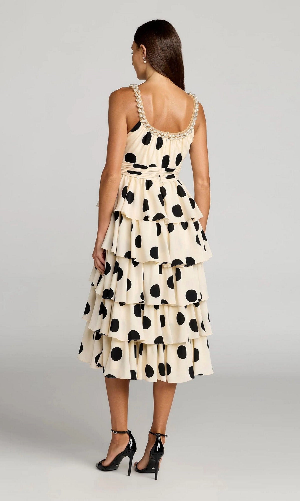 A woman stands with her back to the camera, wearing the Mac Duggal 27736 Midi Party Dress—an ivory, tiered party dress adorned with large black polka dots—paired with black high heels.