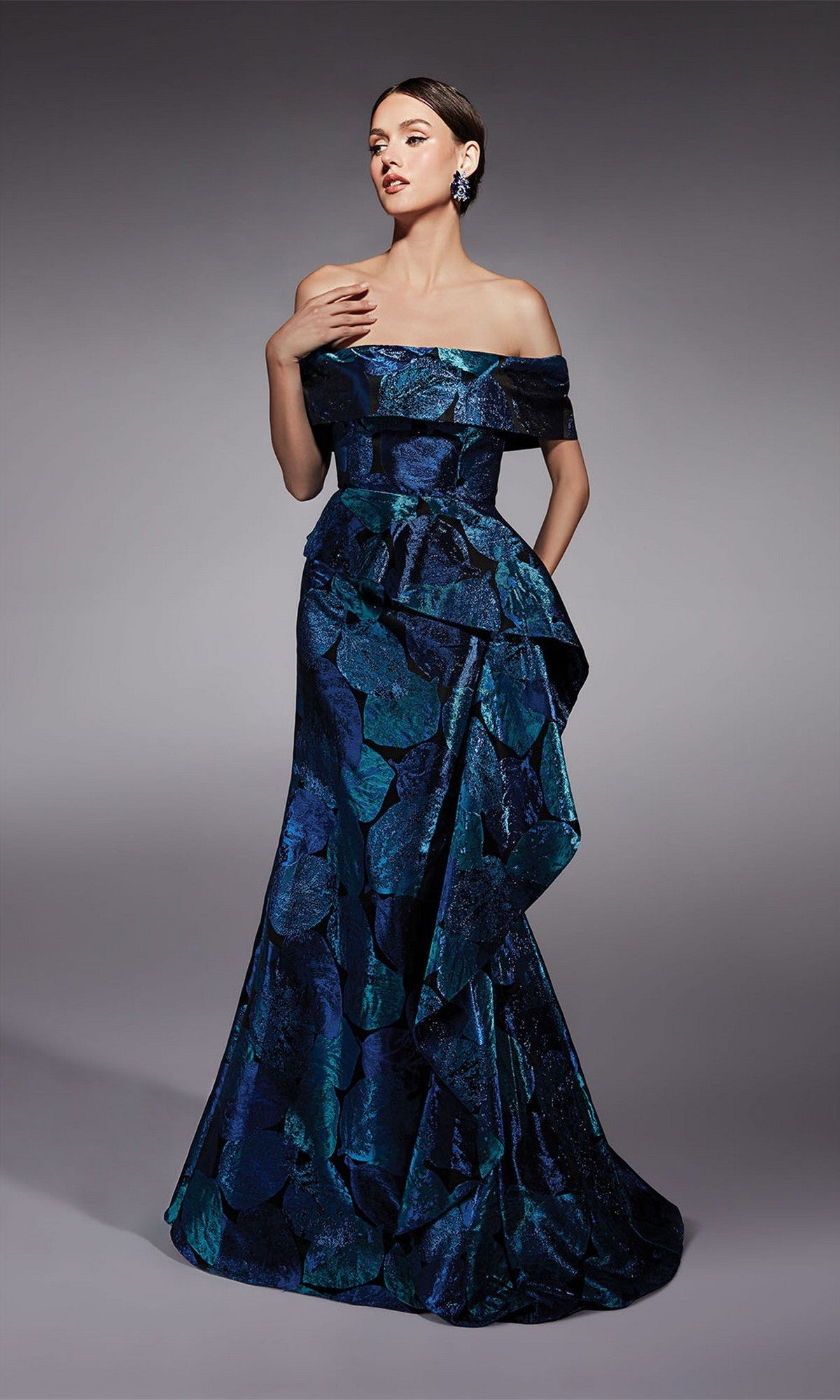 A woman stands against a plain gray background wearing the Alyce 27736 long formal dress in royal, featuring an off-the-shoulder neckline, ruffle details, and a fit-and-flare silhouette.