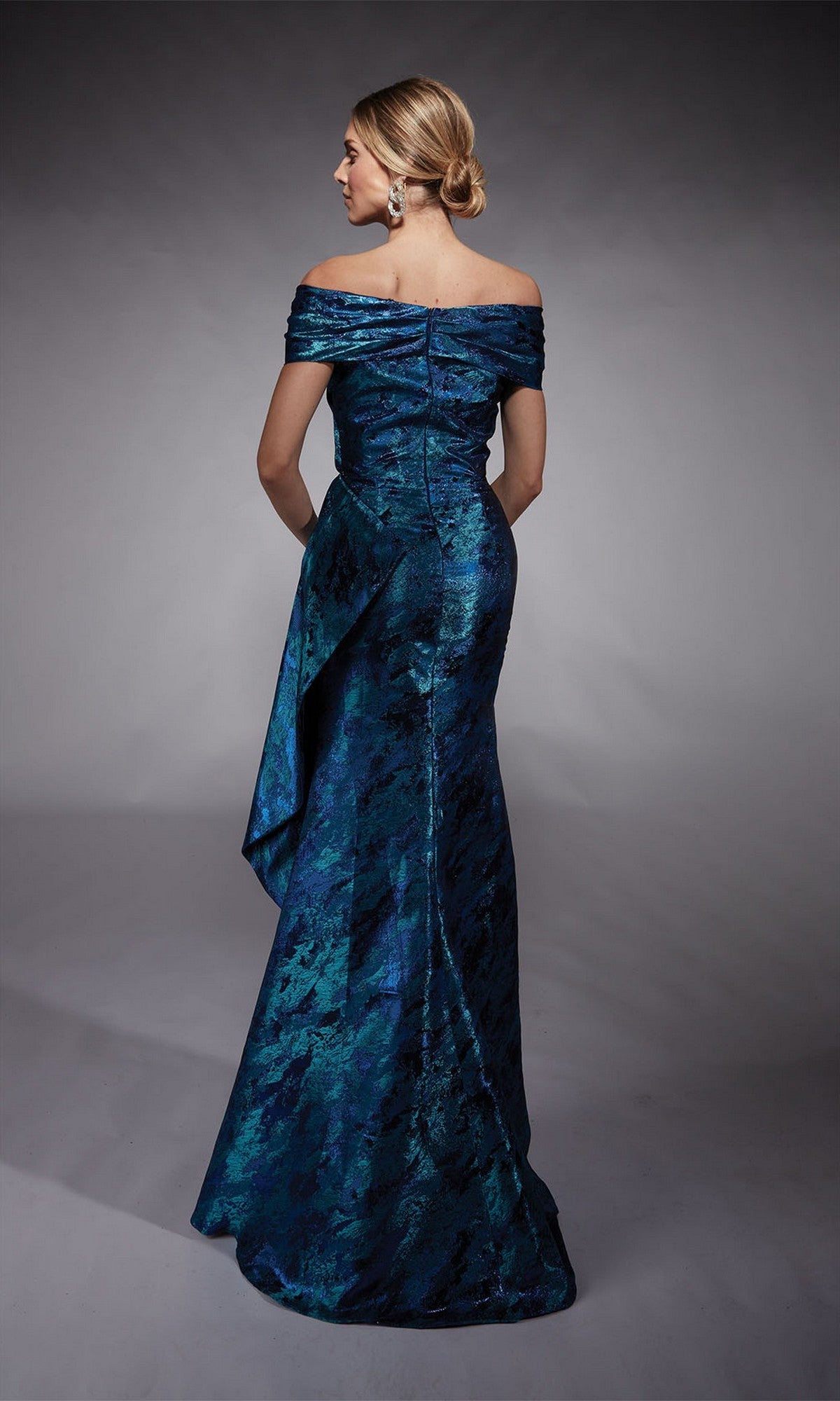 A woman stands with her back to the camera in the Alyce 27737 long formal dress, featuring an off-the-shoulder blue and green jacquard design—a refined mother-of-the-bride choice against a gray backdrop.