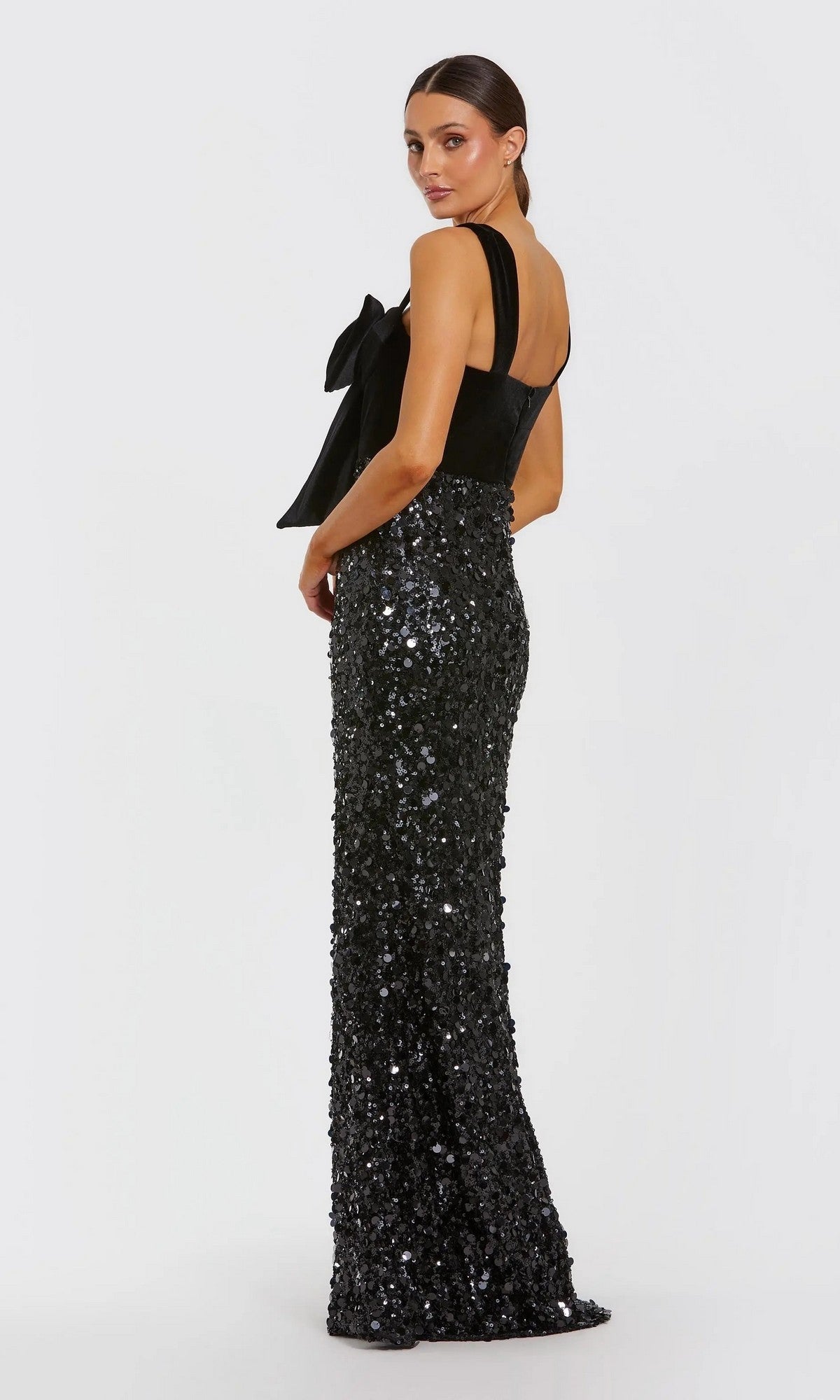 Model wearing the Mac Duggal 27738 long formal dress with a black velvet bodice, large shoulder bow, and sequined floor-length skirt, posed against a plain white background.