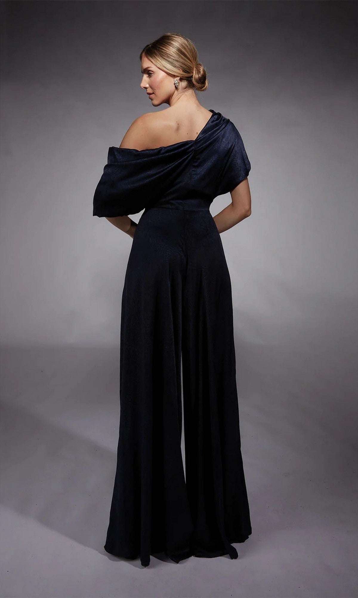 A woman faces away, wearing the Long Formal Jumpsuit: Alyce 27743 with off-the-shoulder style and wide legs—an elegant mother-of-the-bride outfit—set against a plain gray background.