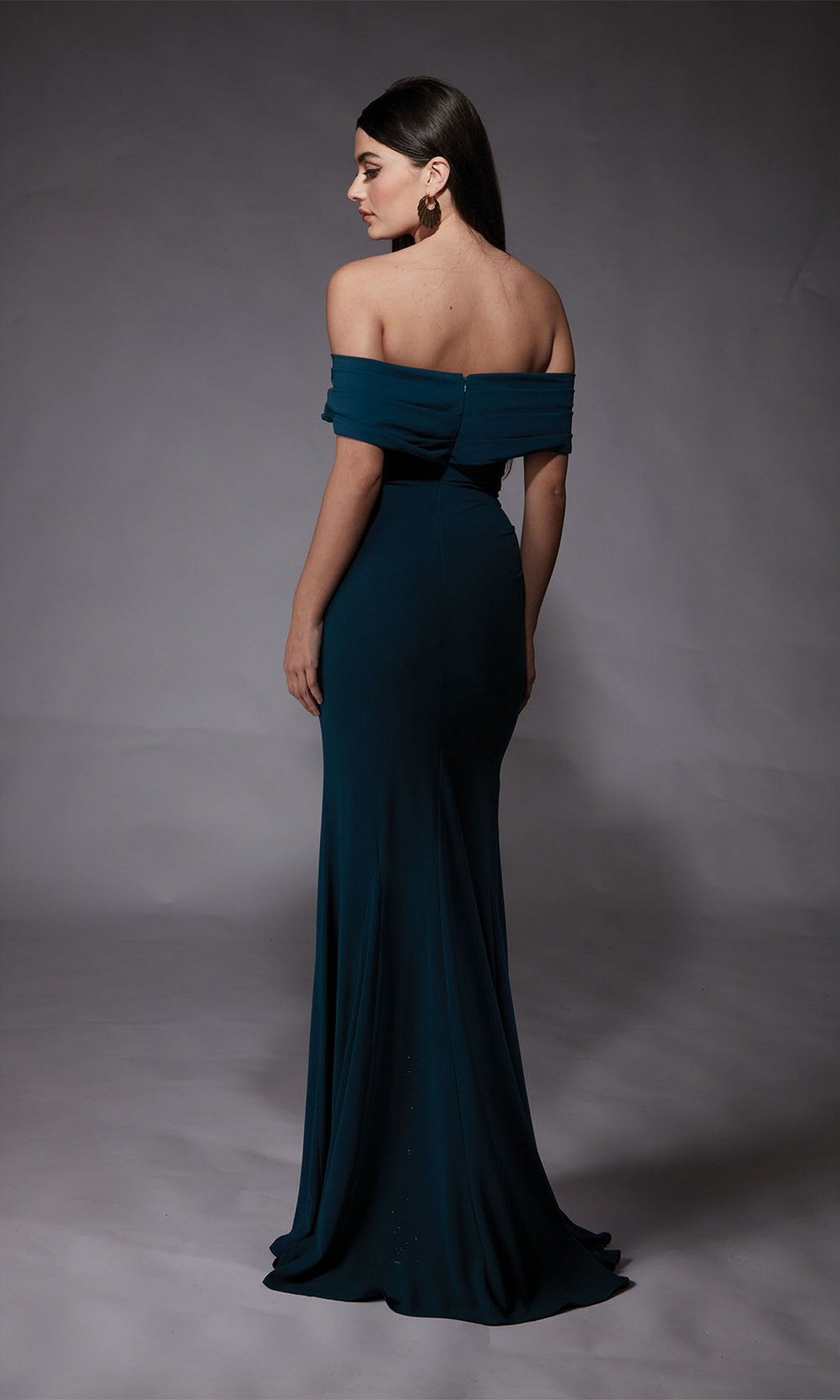 A woman stands facing away in the Long Formal Dress: Alyce 27745, highlighting the elegant back and side of the dark teal off-the-shoulder gown—perfect for prom—against a plain gray backdrop.