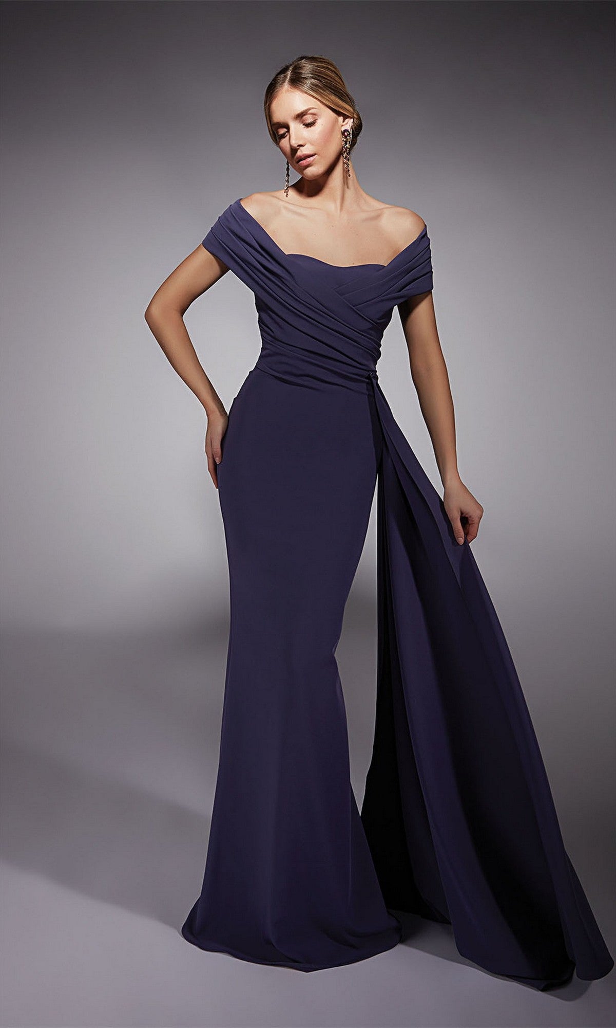 A woman stands in a studio wearing the Alyce 27745 long formal dress in grape, featuring an off-the-shoulder design with draped details and a side sash—ideal for prom.