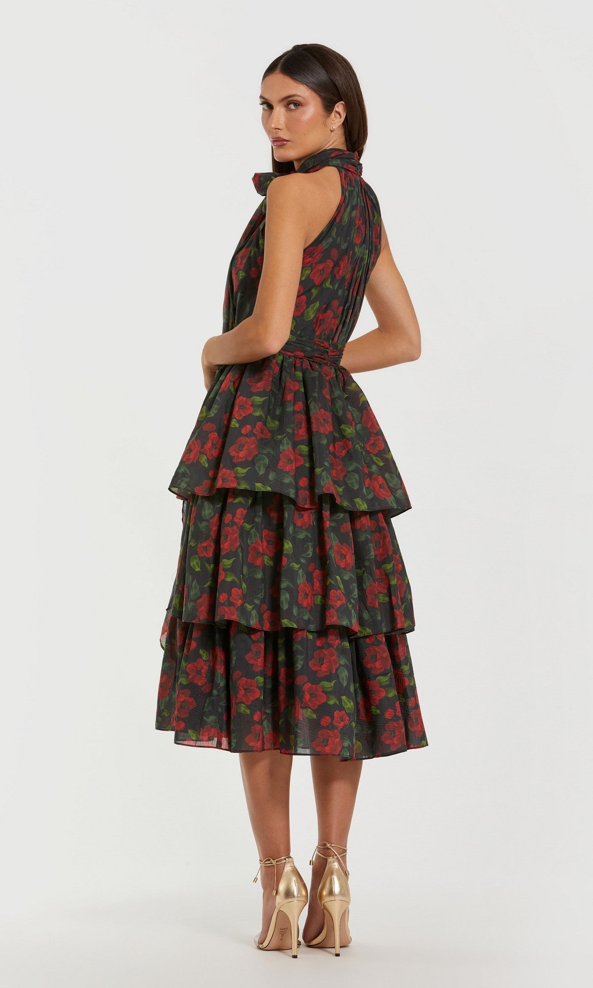 A woman models the Mac Duggal 27747 Midi Wedding Guest Dress, a sleeveless, tiered tea-length design with a red floral print on dark fabric, paired with gold heels and set against a plain white background.