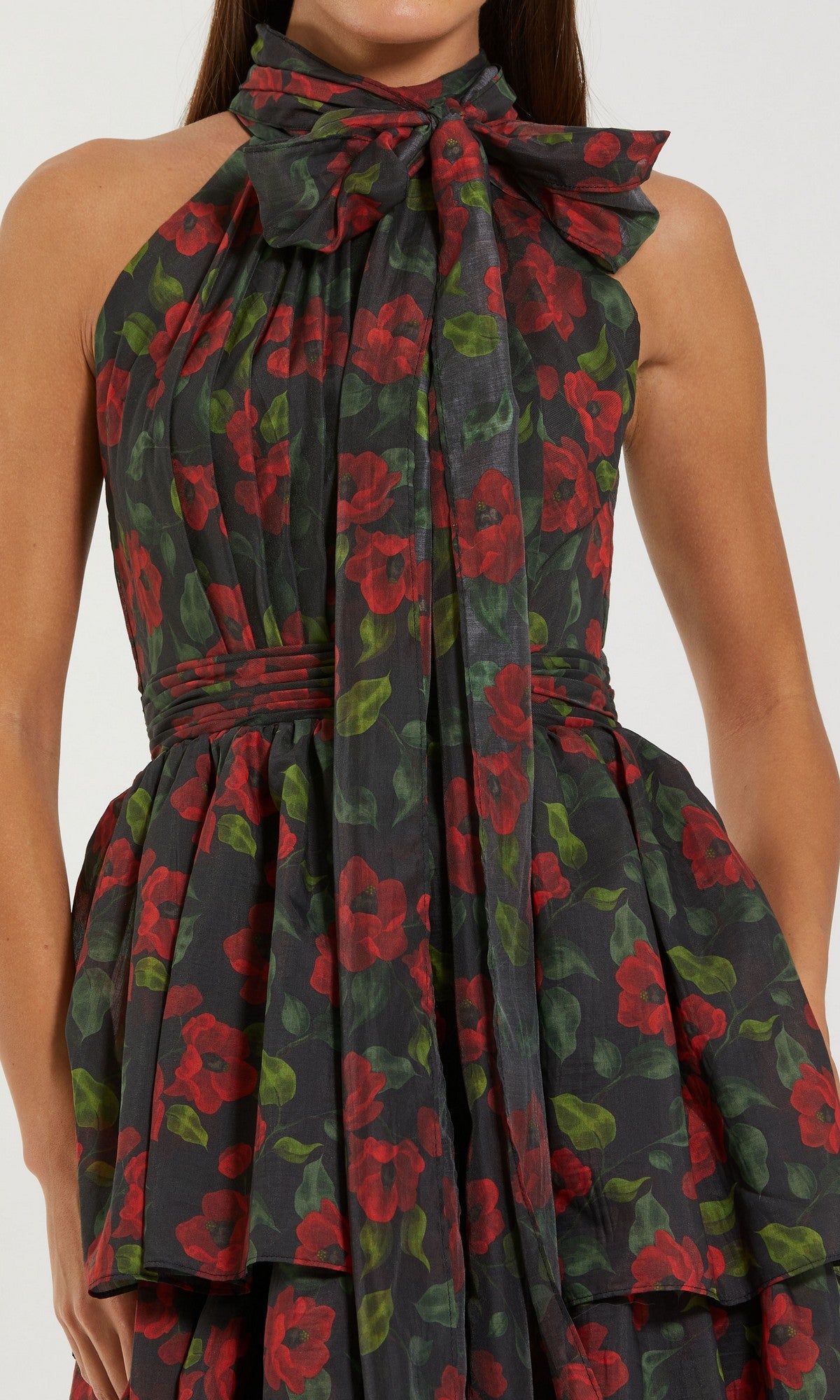 A woman in the Mac Duggal 27747 midi wedding guest dress featuring a sleeveless black design with red and green floral print, layered skirt, and a large bow at the neckline.