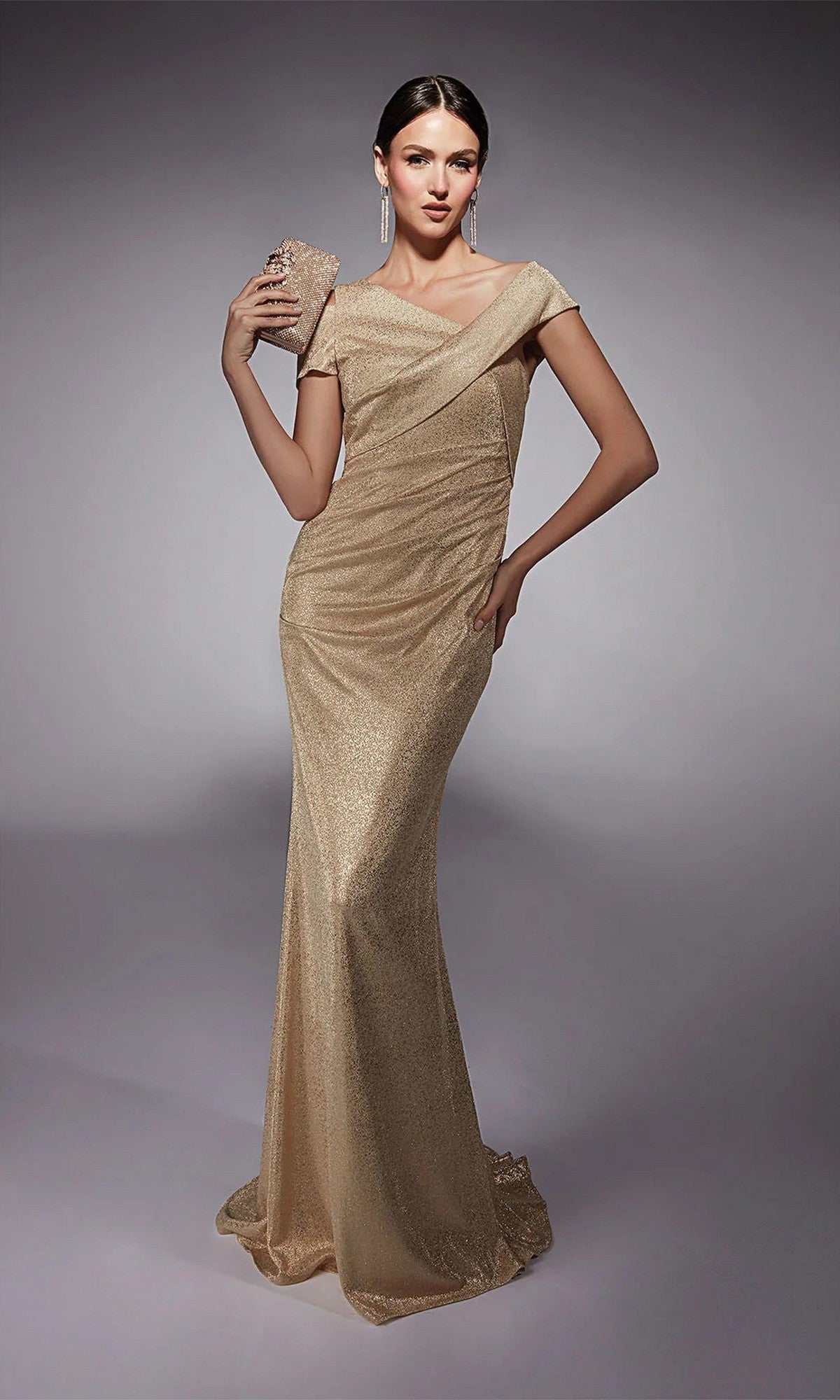 A woman models the Alyce 27762 long formal dress in gold, featuring an off-the-shoulder glitter jersey design. She poses against a neutral background with a matching clutch and long drop earrings.