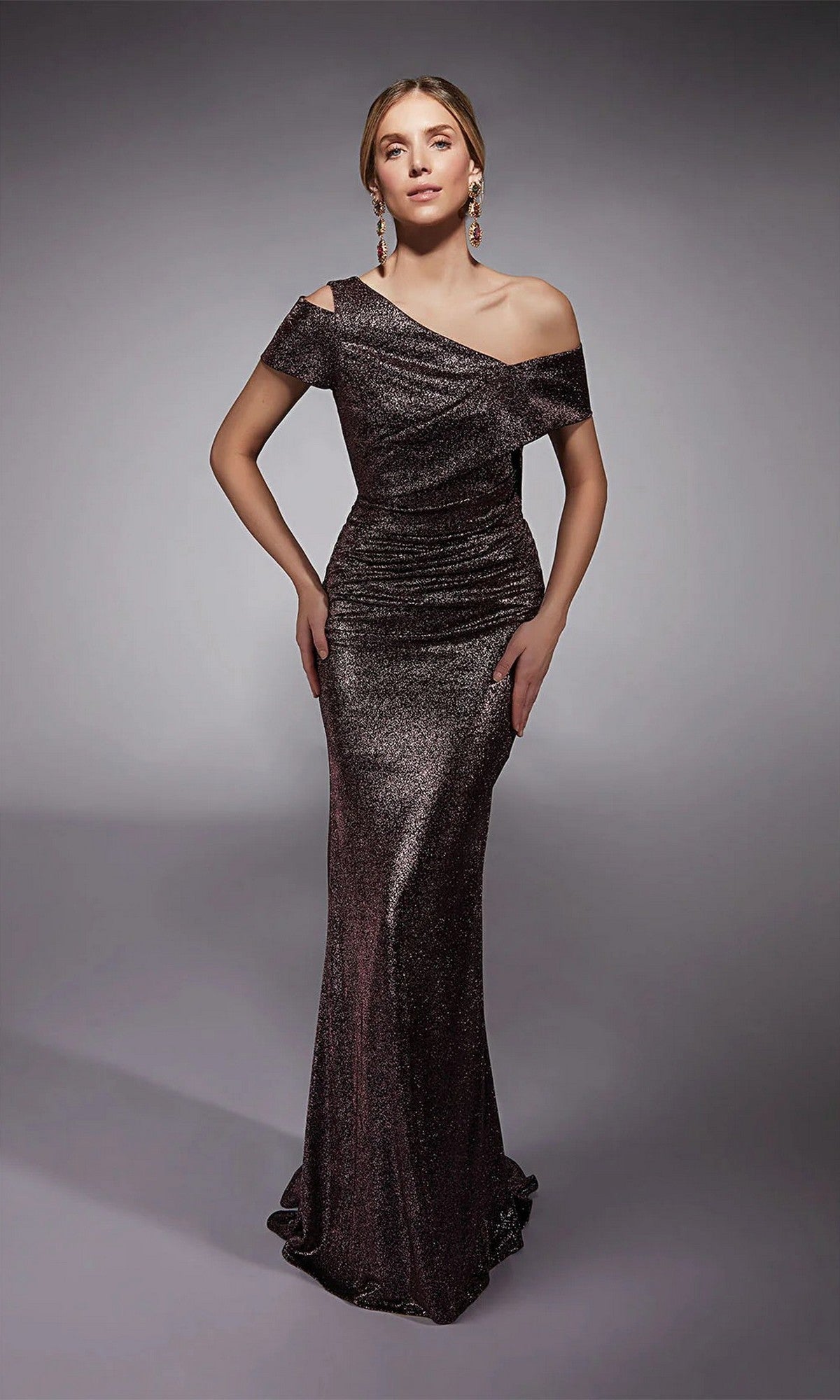 A woman poses in a studio wearing the Alyce 27762 long formal dress in dark mauve, featuring a shimmering glitter jersey fabric, off-the-shoulder neckline, fitted silhouette, and subtle ruched detailing.