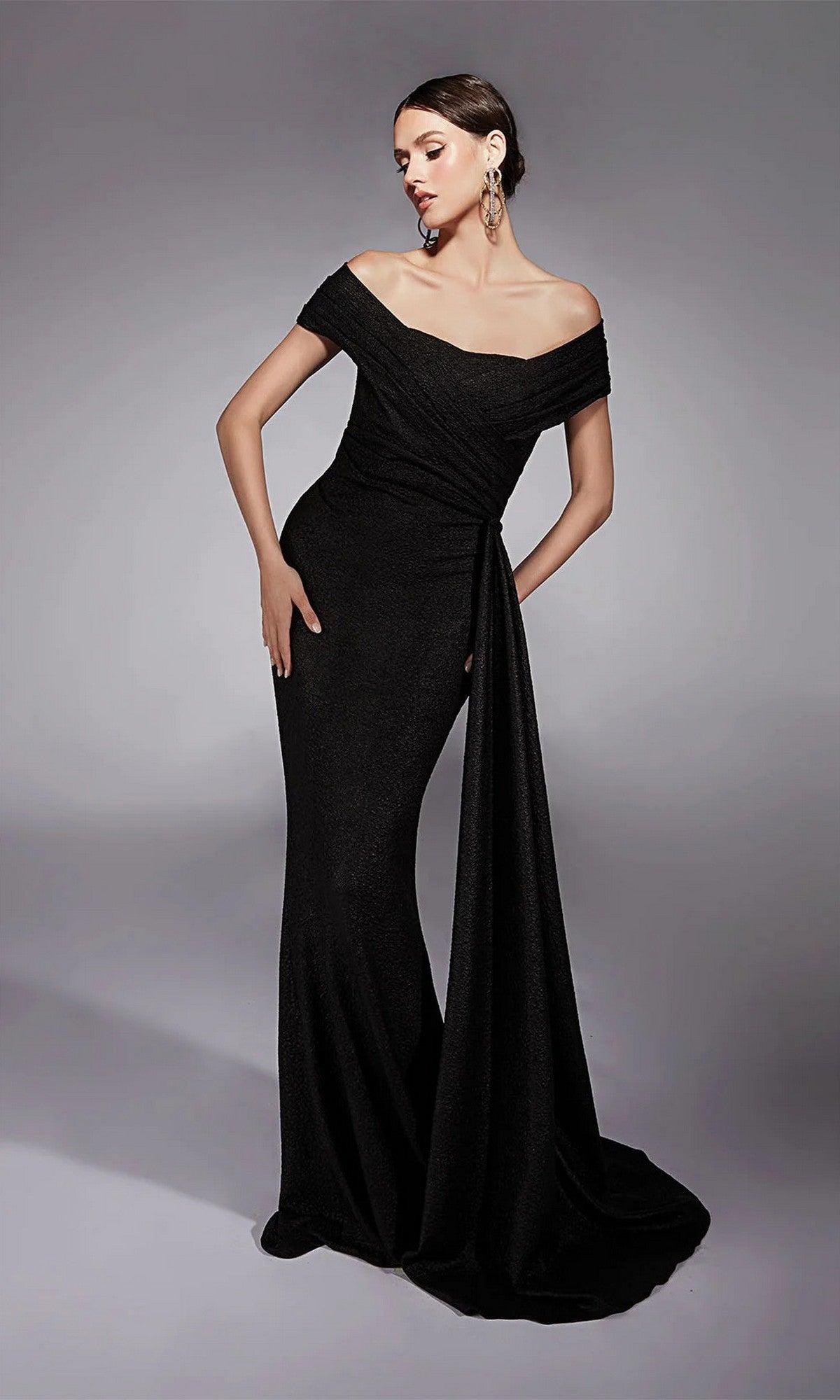 A woman poses in the Alyce 27767, a black, off-the-shoulder long formal dress with a draped sash and floor-length hem—ideal as a mother-of-the-bride gown—against a plain gray background.
