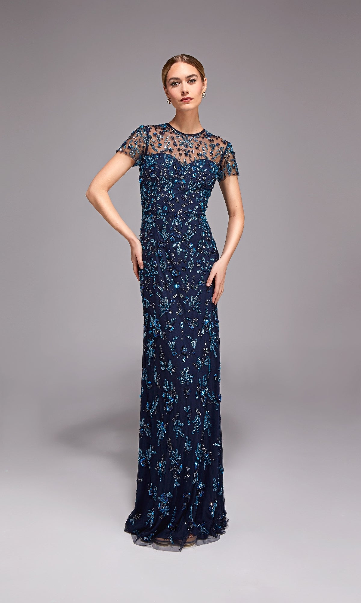 A woman wears the Alyce 27789 long formal dress in navy blue with short sleeves and blue floral embroidery, standing against a plain gray background.