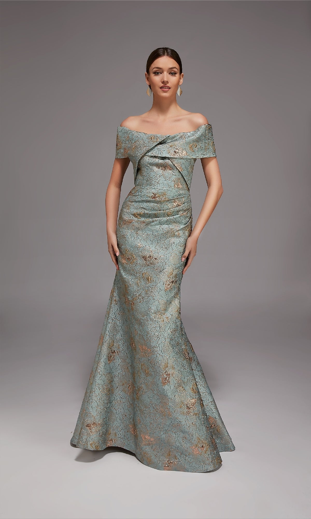 A woman stands against a plain gray background wearing the Alyce 27802 Long Formal Dress, an off-the-shoulder, floor-length gown featuring a blue and gold brocade pattern.