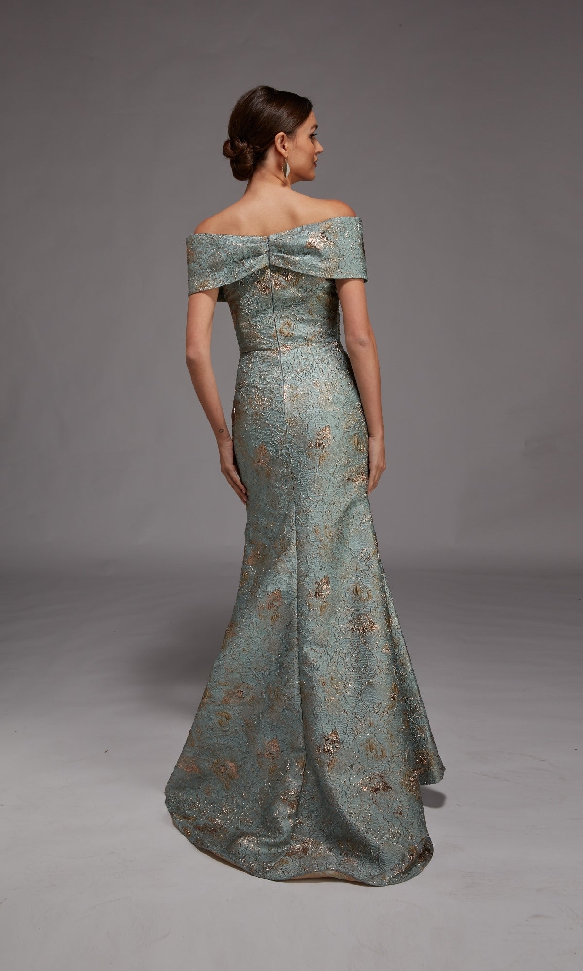 A woman faces away, wearing the Alyce 27802 Long Formal Dress—an off-the-shoulder, floor-length gown with a fitted bodice and metallic floral pattern—set against a plain gray background.