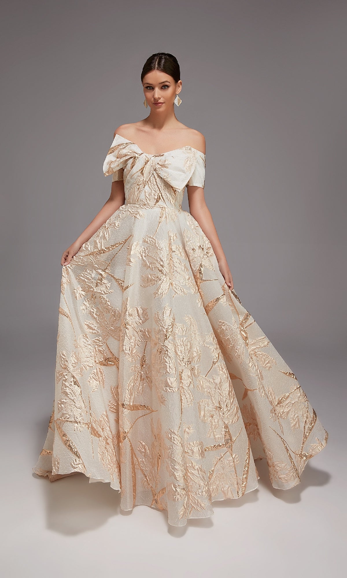 A woman poses in a studio wearing the Alyce 27804 long formal dress, an off-the-shoulder ball gown with a large bow and gold floral jacquard patterns.