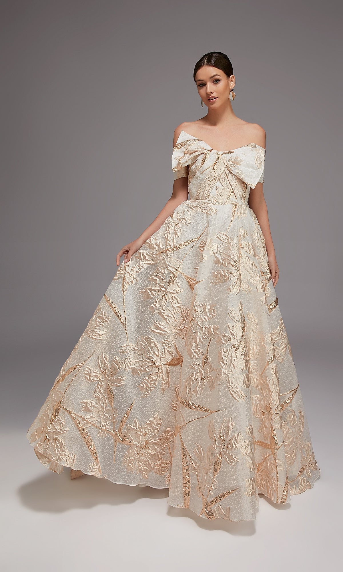 A woman stands against a gray background wearing the Alyce 27804, an ivory/gold off-the-shoulder, floor-length formal dress in luxurious jacquard with a large bow and gold floral patterns.