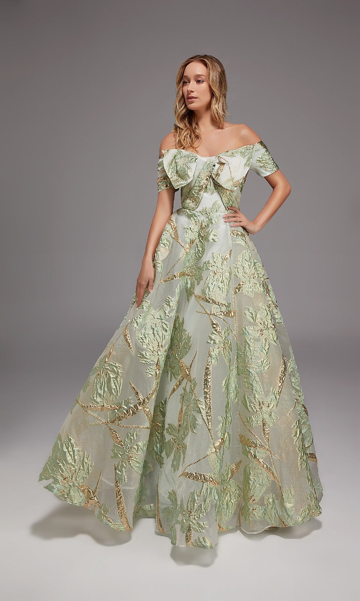 In a studio, a woman wears the Alyce 27804 long formal dress in Sea Glass/Gold, featuring an off-the-shoulder design and floor-length silhouette adorned with metallic leaf motifs.
