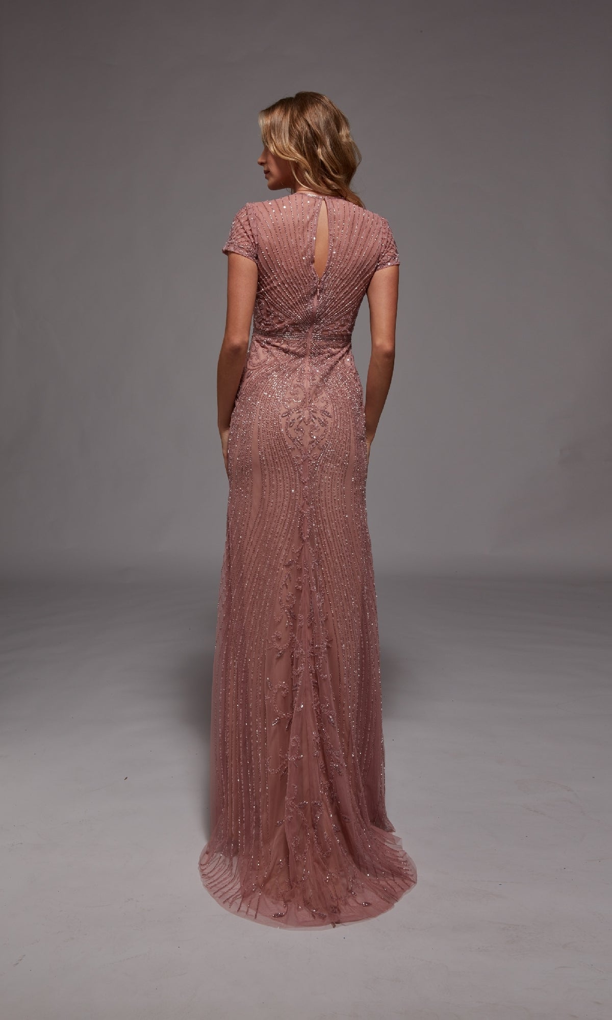 A woman in the Alyce 27823 long pink beaded evening dress with short sleeves stands facing away against a plain gray background.