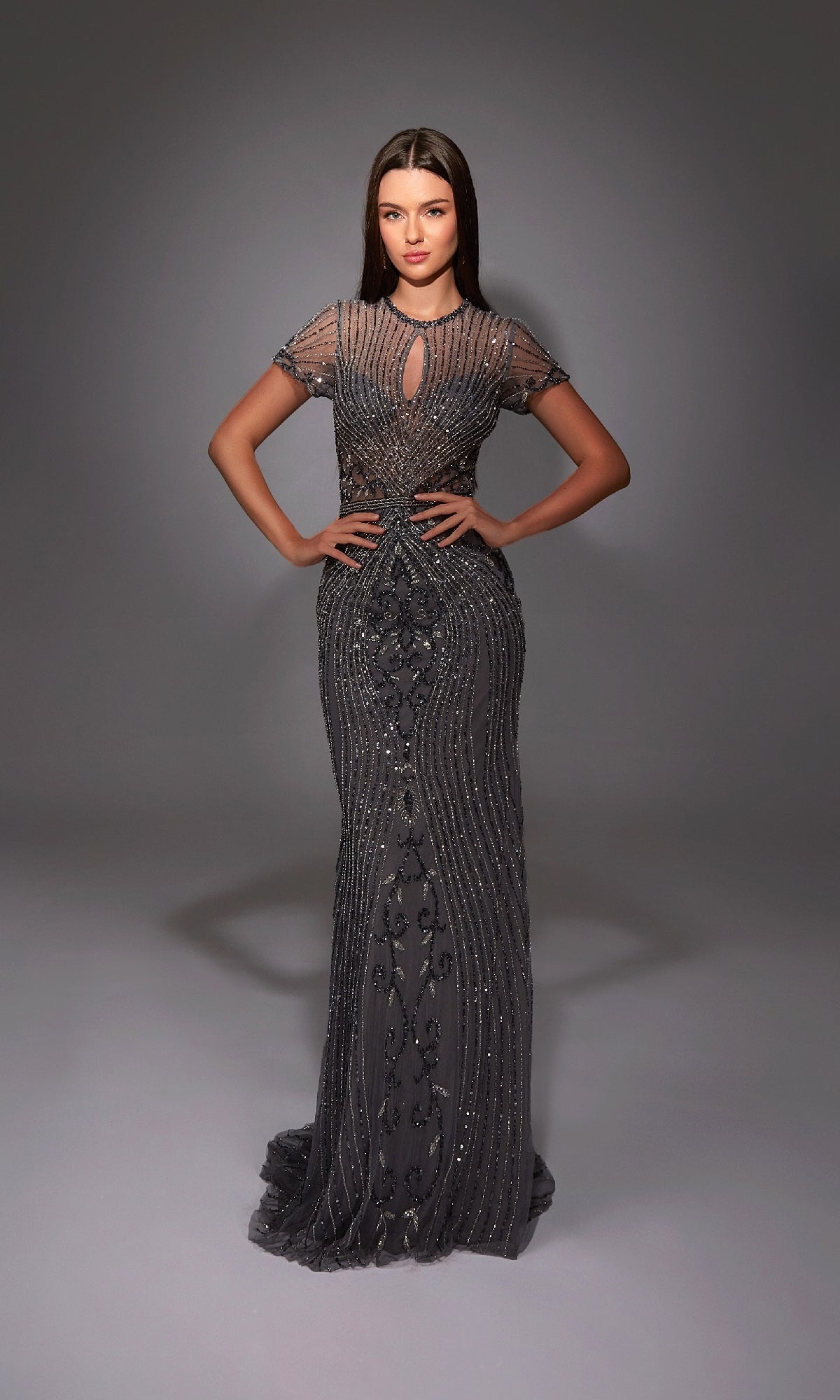 A woman stands with hands on hips, wearing the Alyce 27823 long formal dress—a dark, beaded gown with short sleeves and a keyhole neckline—against a gray studio backdrop.