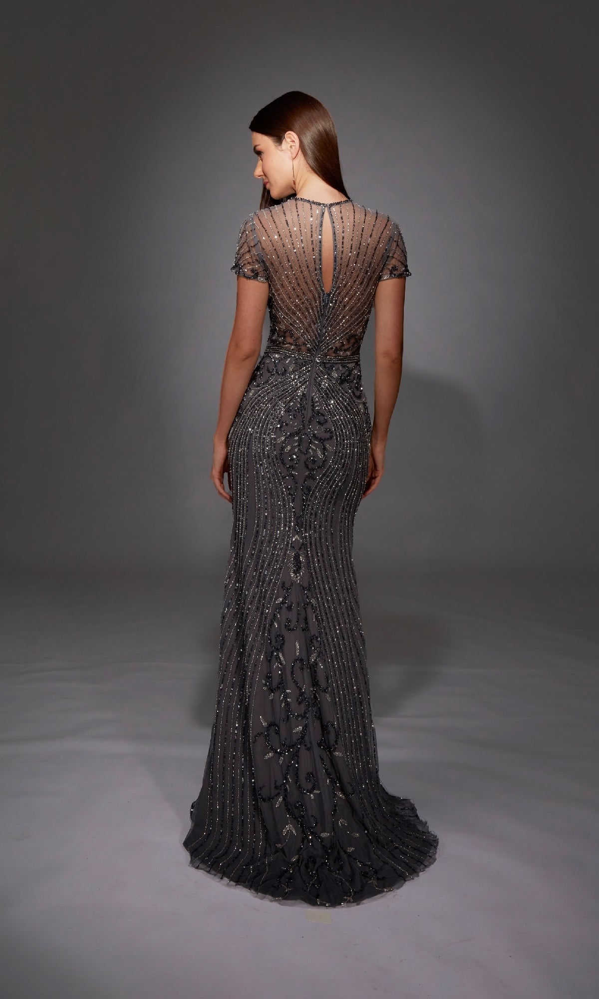 A woman faces away, wearing the Alyce 27823 long formal dress—a sheer black gown with intricate beadwork and a keyhole back—set against a plain gray background.