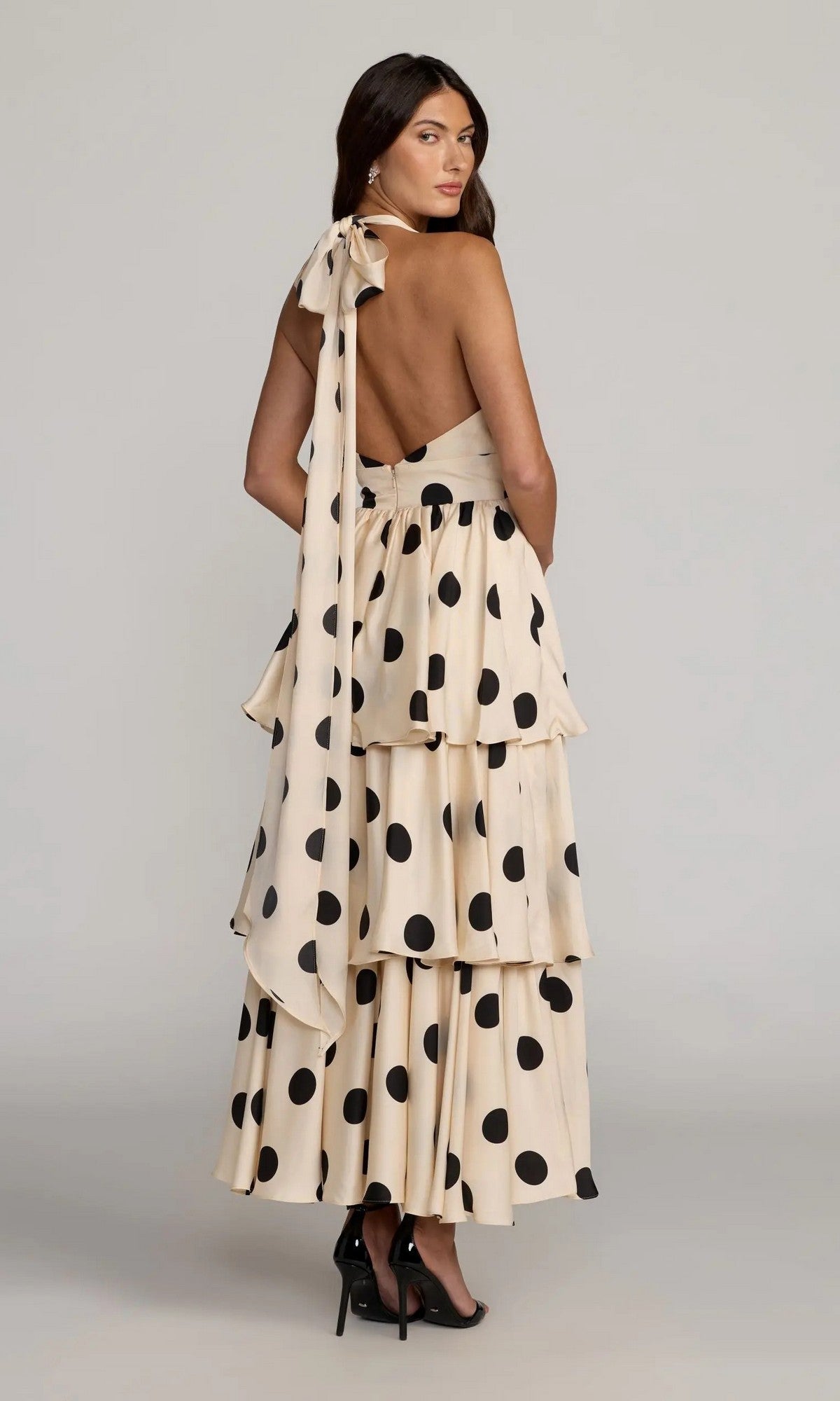 A woman stands in black heels against a plain background, wearing the Mac Duggal 27833 long formal dress—a tiered, backless cream midi dress adorned with large black polka dots and finished with a neck bow.