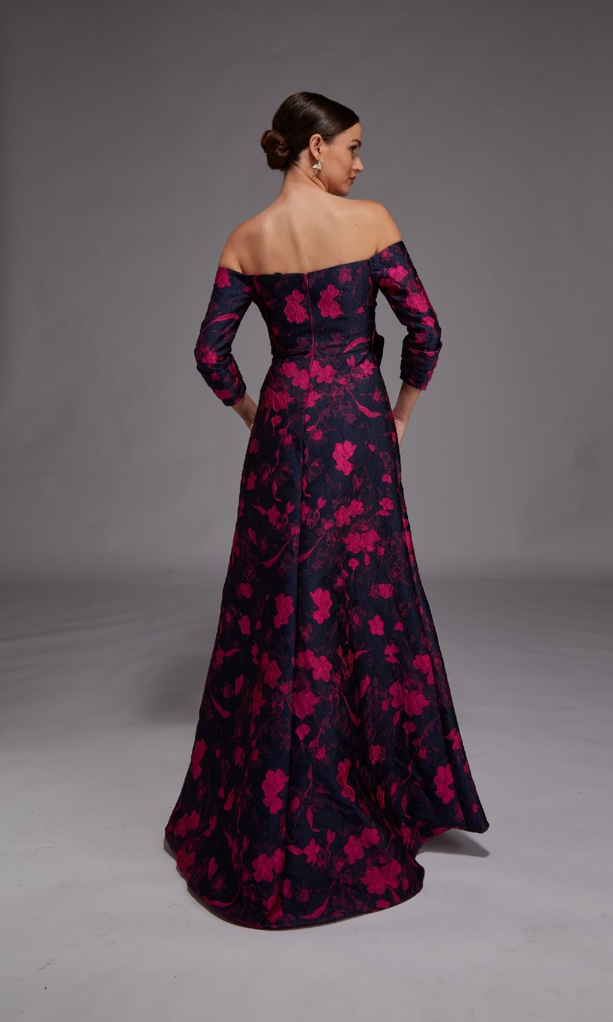 A woman faces away from the camera in the Alyce 27833, a long formal dress with an off-the-shoulder, floor-length A-line silhouette in dark fabric adorned with pink floral patterns.