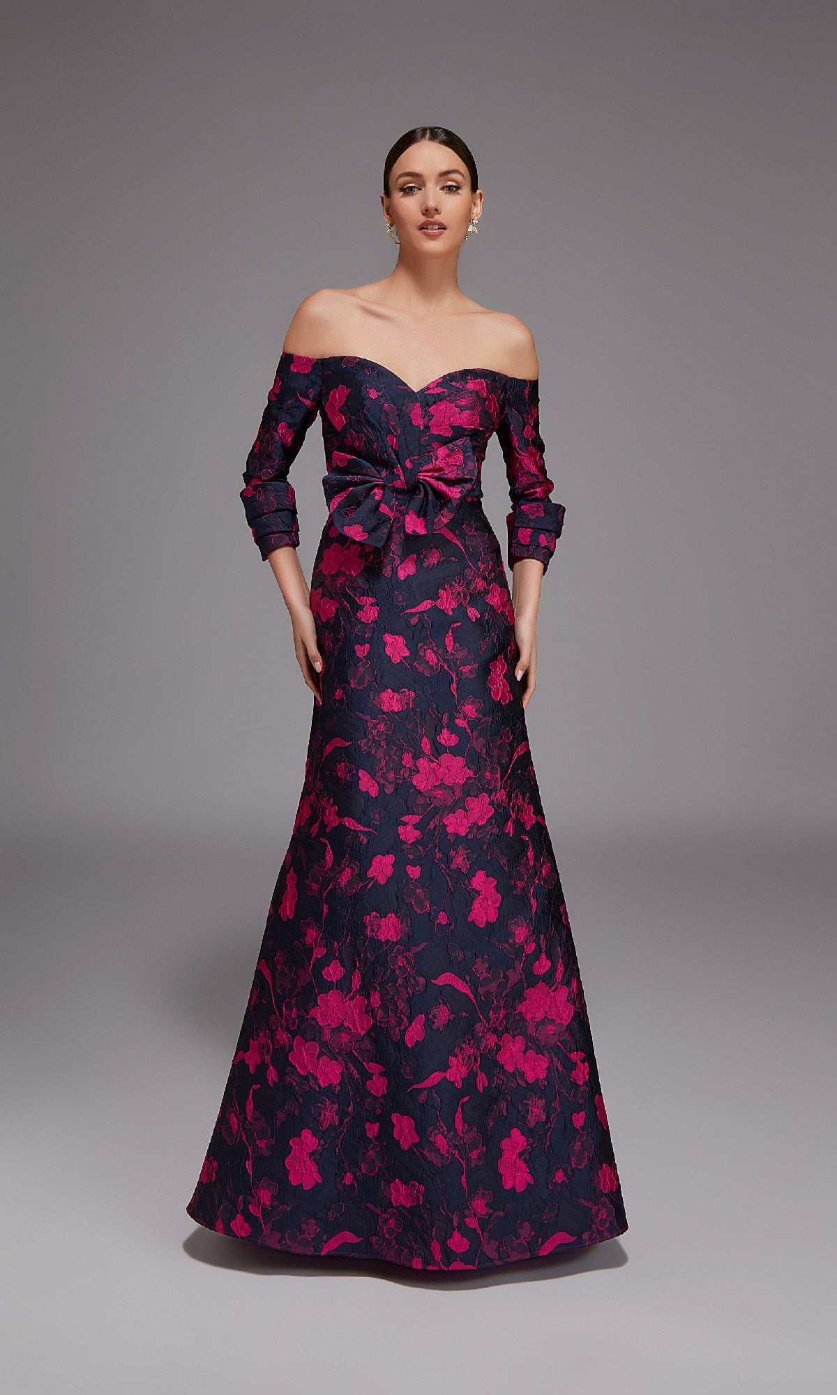 A woman wears the Alyce 27833 long formal dress, an off-the-shoulder jacquard gown with a dark base, bold pink floral print, fitted bodice, waist bow, A-line silhouette, and graceful floor-length hem.